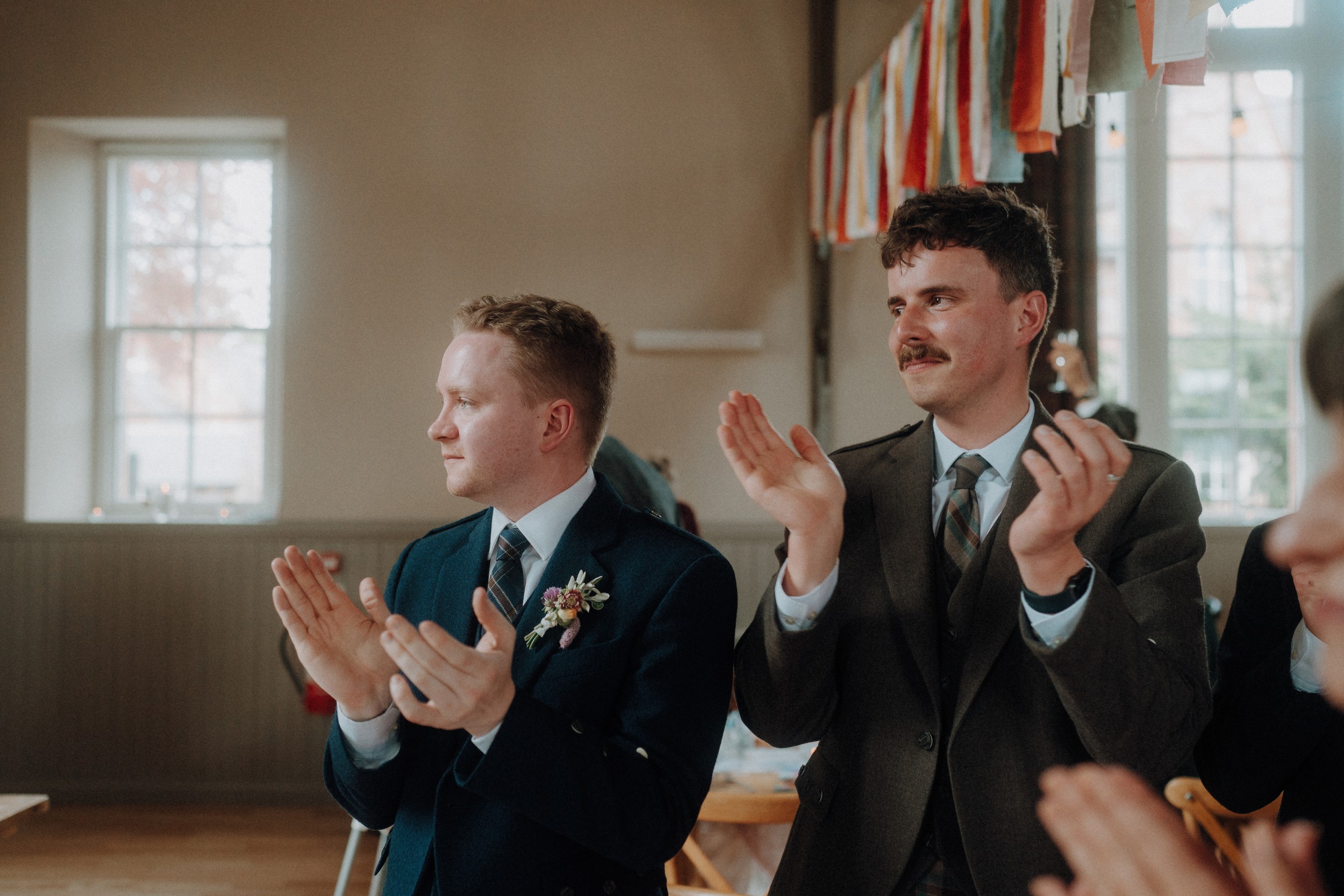Two men in suits sit indoors, clapping and looking to the left. Colorful fabric streamers hang above them; daylight comes through the windows in the background.