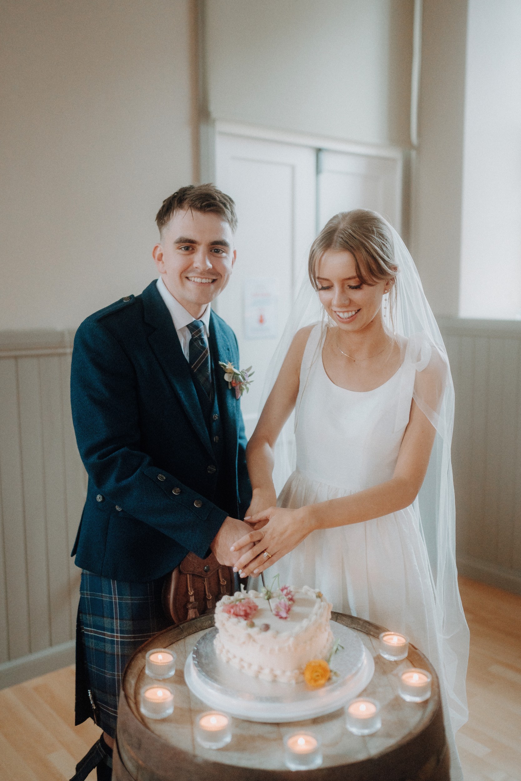 A bride and groom, dressed in wedding attire, smile while cutting a small floral cake together, surrounded by tealight candles on a round table.
