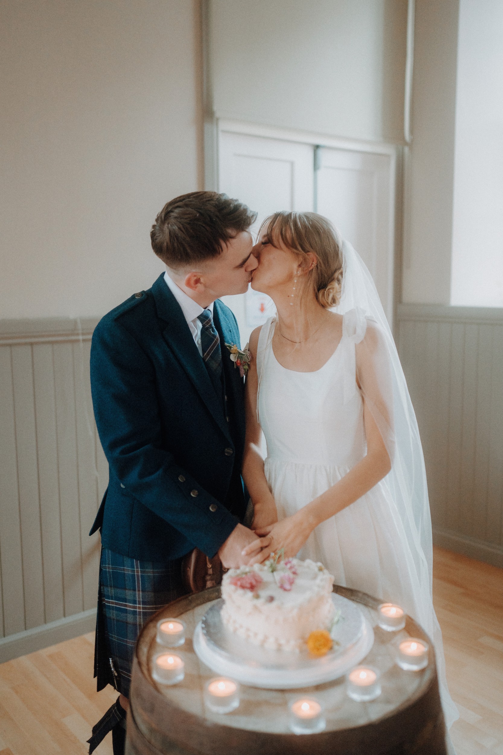 A bride and groom kiss while cutting their wedding cake together, standing in a softly lit room with candles arranged around the cake.