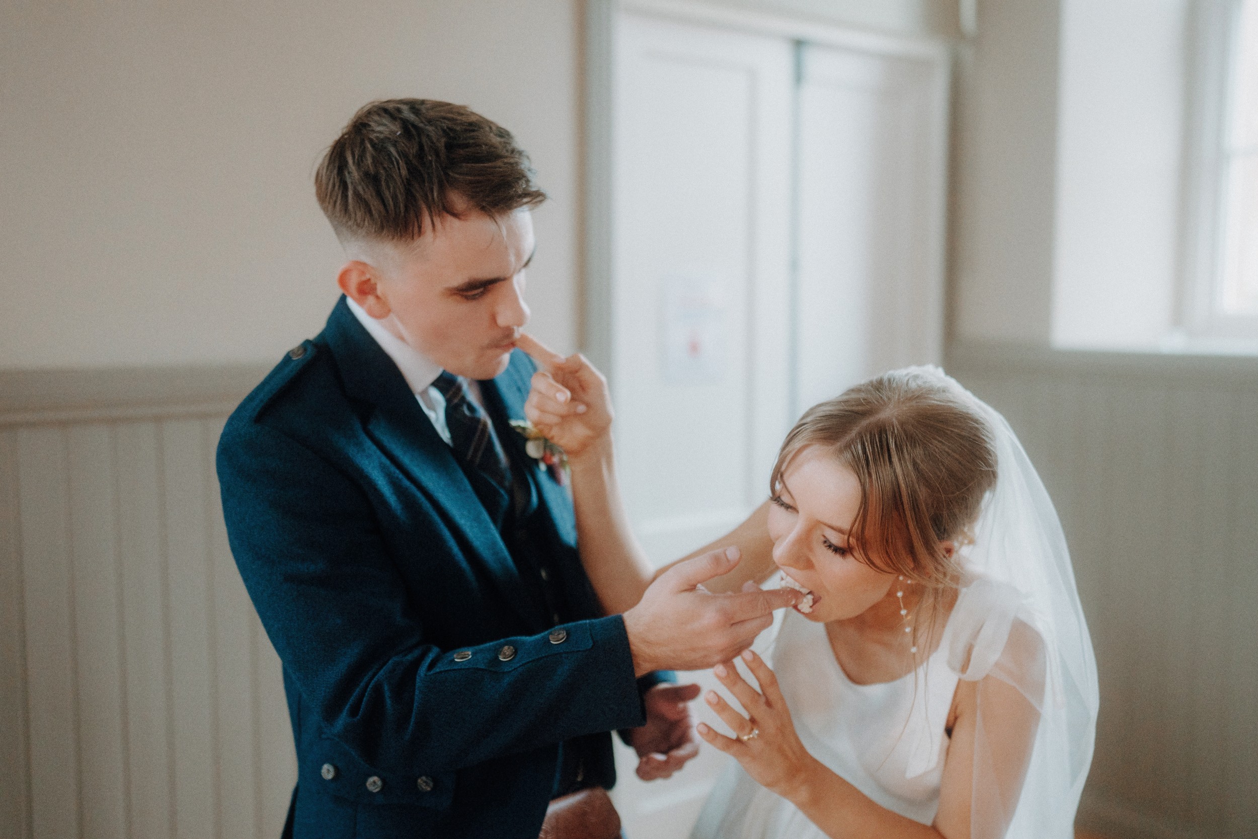 A bride and groom in formal attire feed each other cake in a light-filled room, with the bride wearing a veil and the groom dressed in a dark suit.