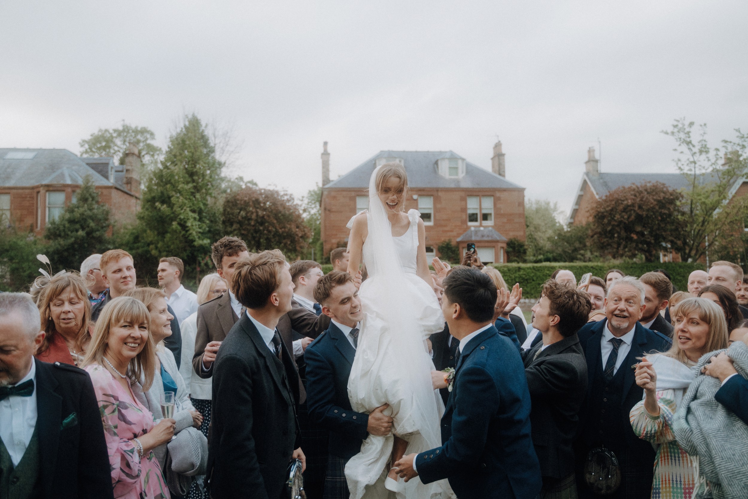 A bride in a white dress is lifted by a group of people outdoors, surrounded by smiling wedding guests in formal attire. Victorian-style houses are visible in the background.