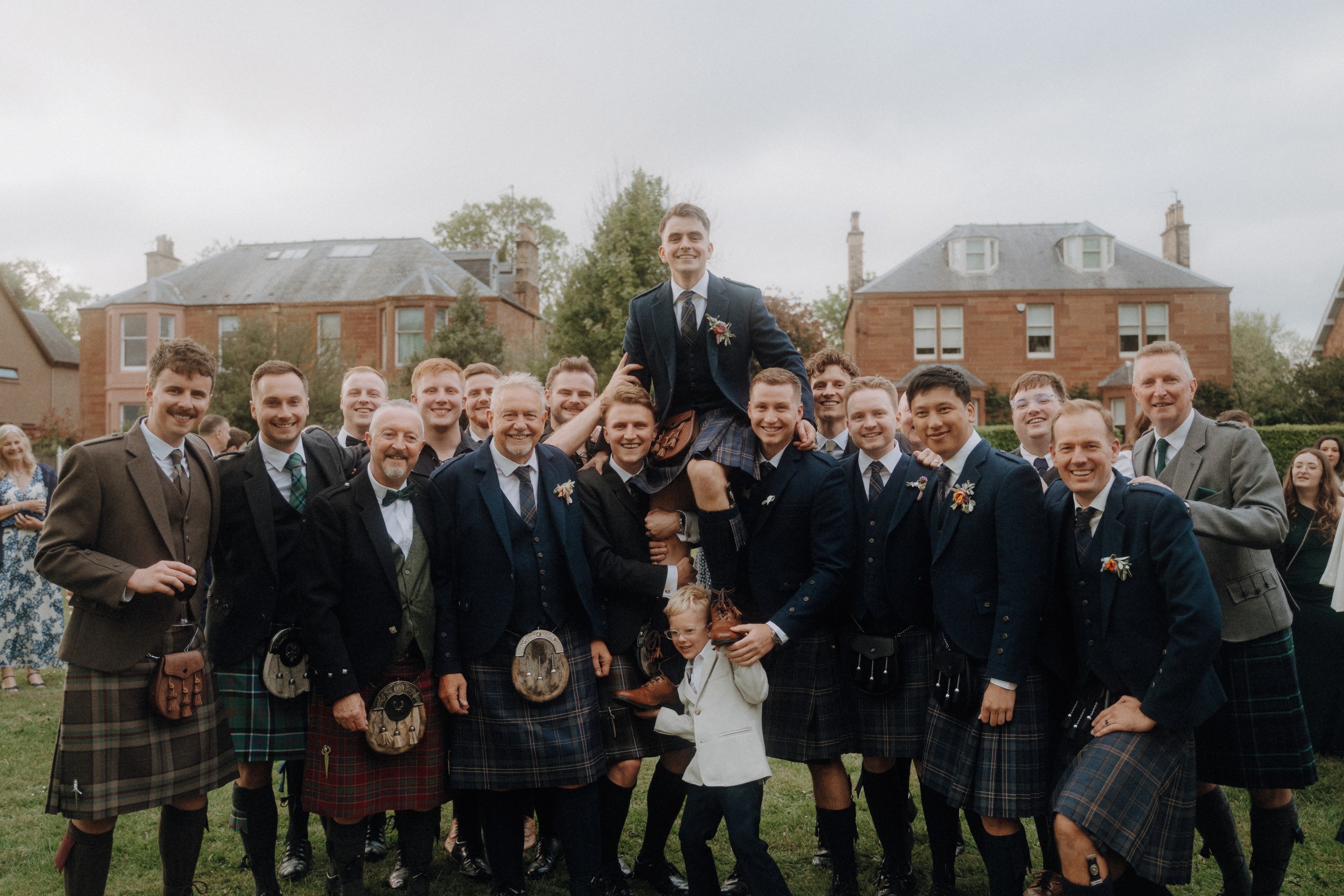 A group of men in kilts and jackets pose outdoors on grass, with one man being lifted on others’ shoulders; houses and trees are visible in the background.