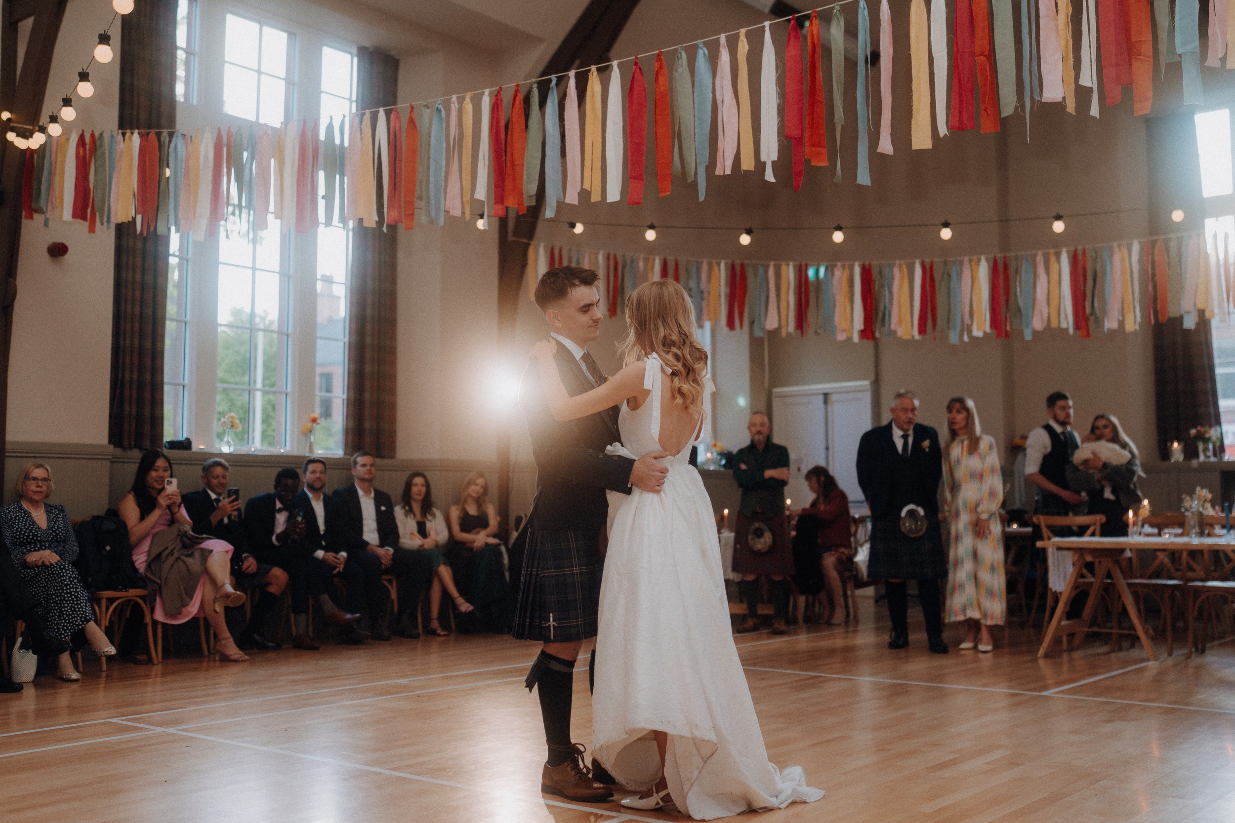 A couple dances in the center of a decorated hall, surrounded by seated and standing guests, with colorful streamers and string lights overhead.