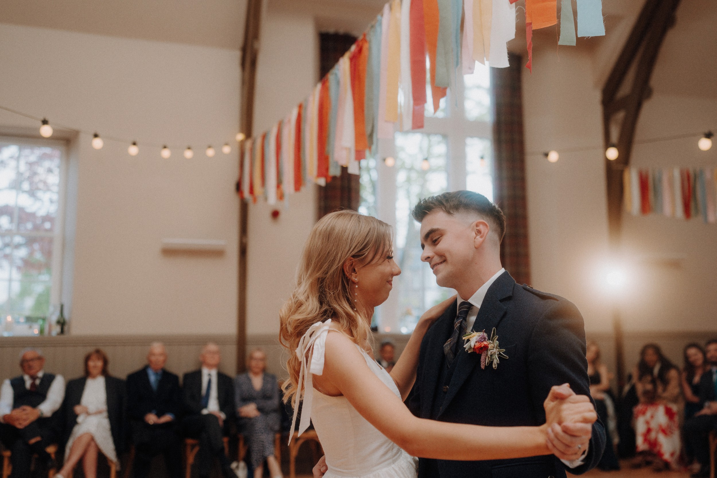 A bride and groom dance together in a decorated hall with guests seated in the background and colorful streamers hanging from the ceiling.