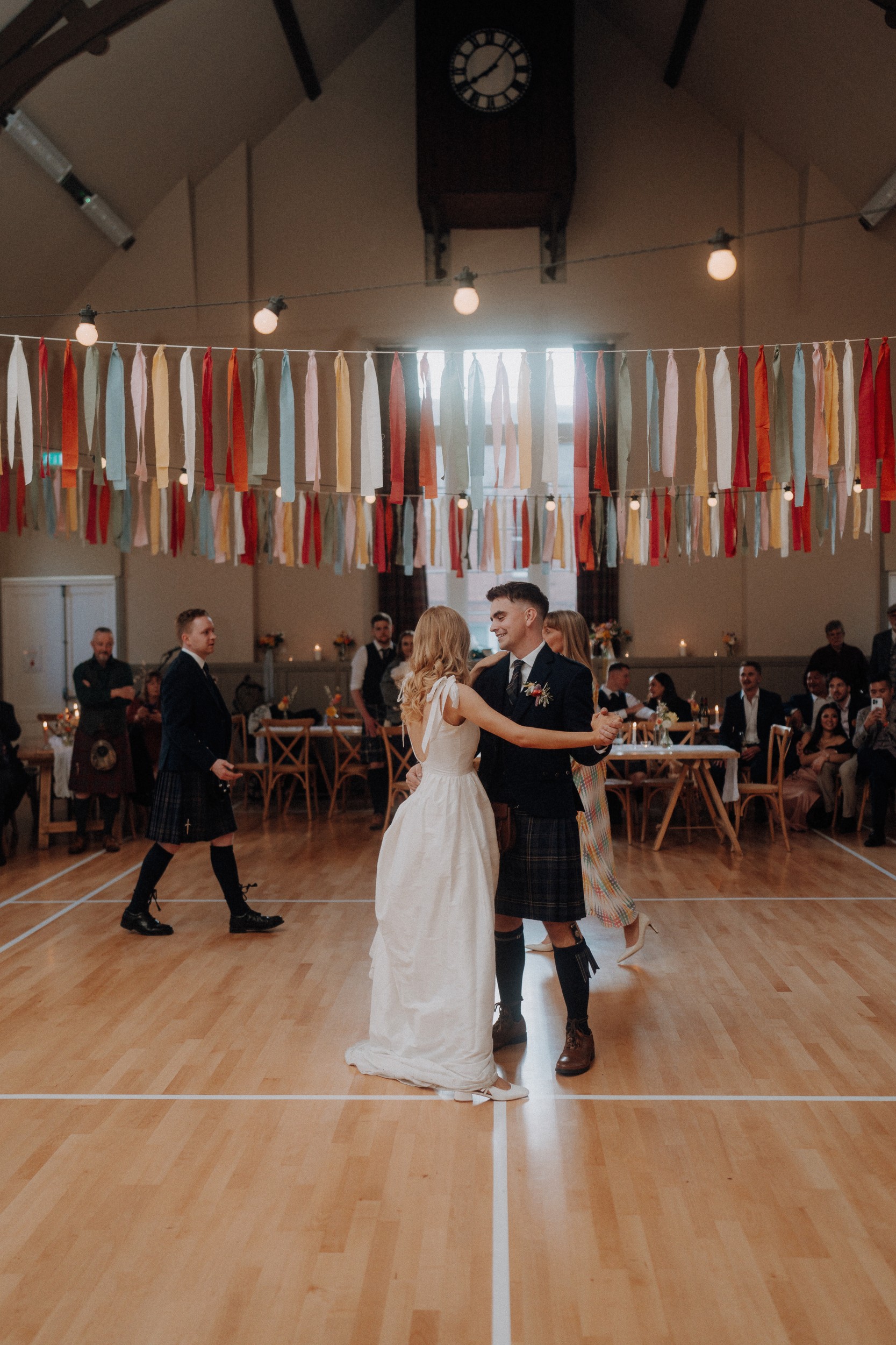 A bride and groom dance in a decorated hall with guests seated around them; colorful streamers hang overhead and a large clock is visible on the wall.