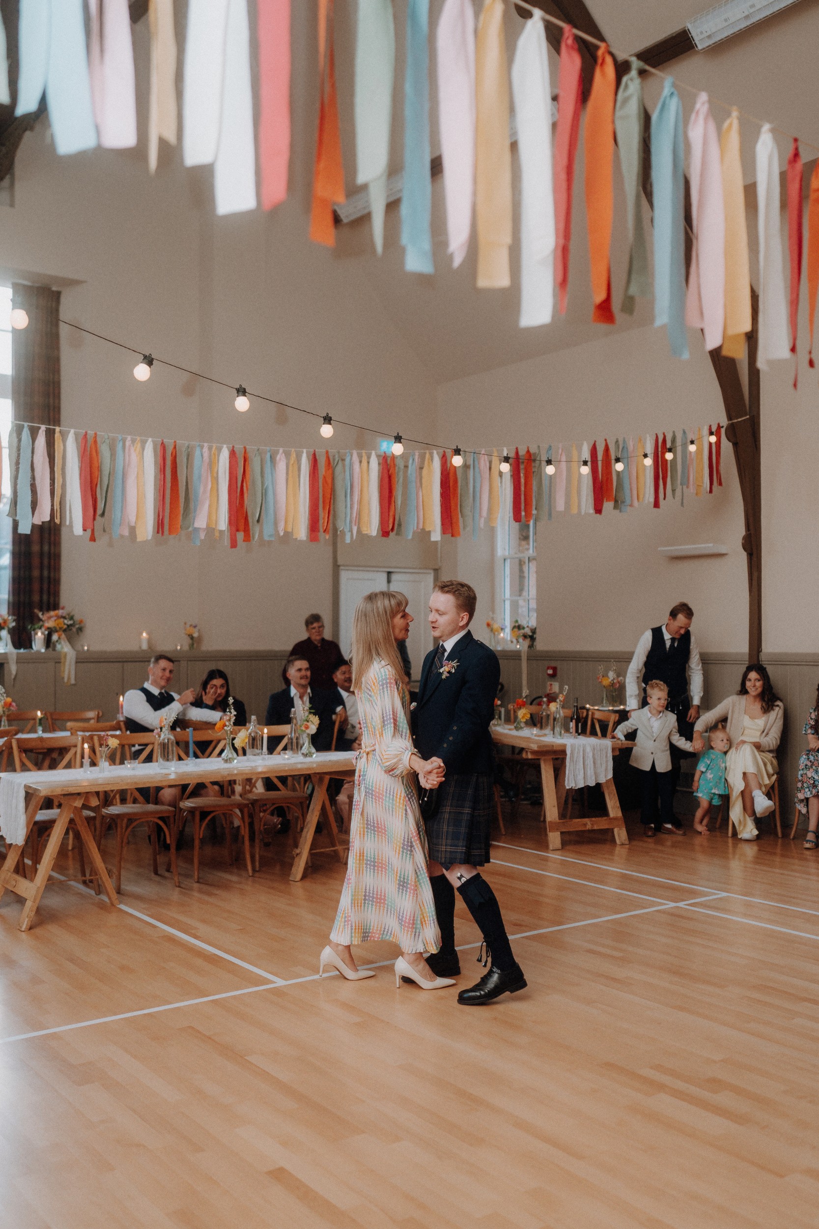 A couple dances in a decorated hall with guests seated at wooden tables, colorful ribbons hanging from the ceiling, and string lights overhead.