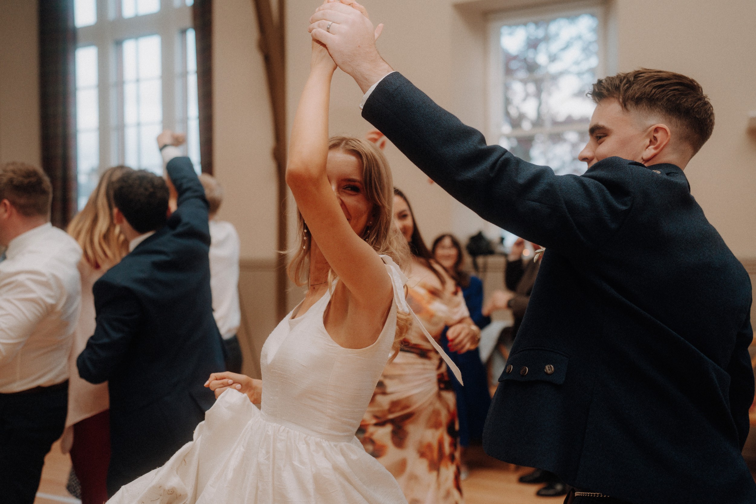A woman in a white dress and a man in a dark suit are dancing together indoors, surrounded by other dancing people.