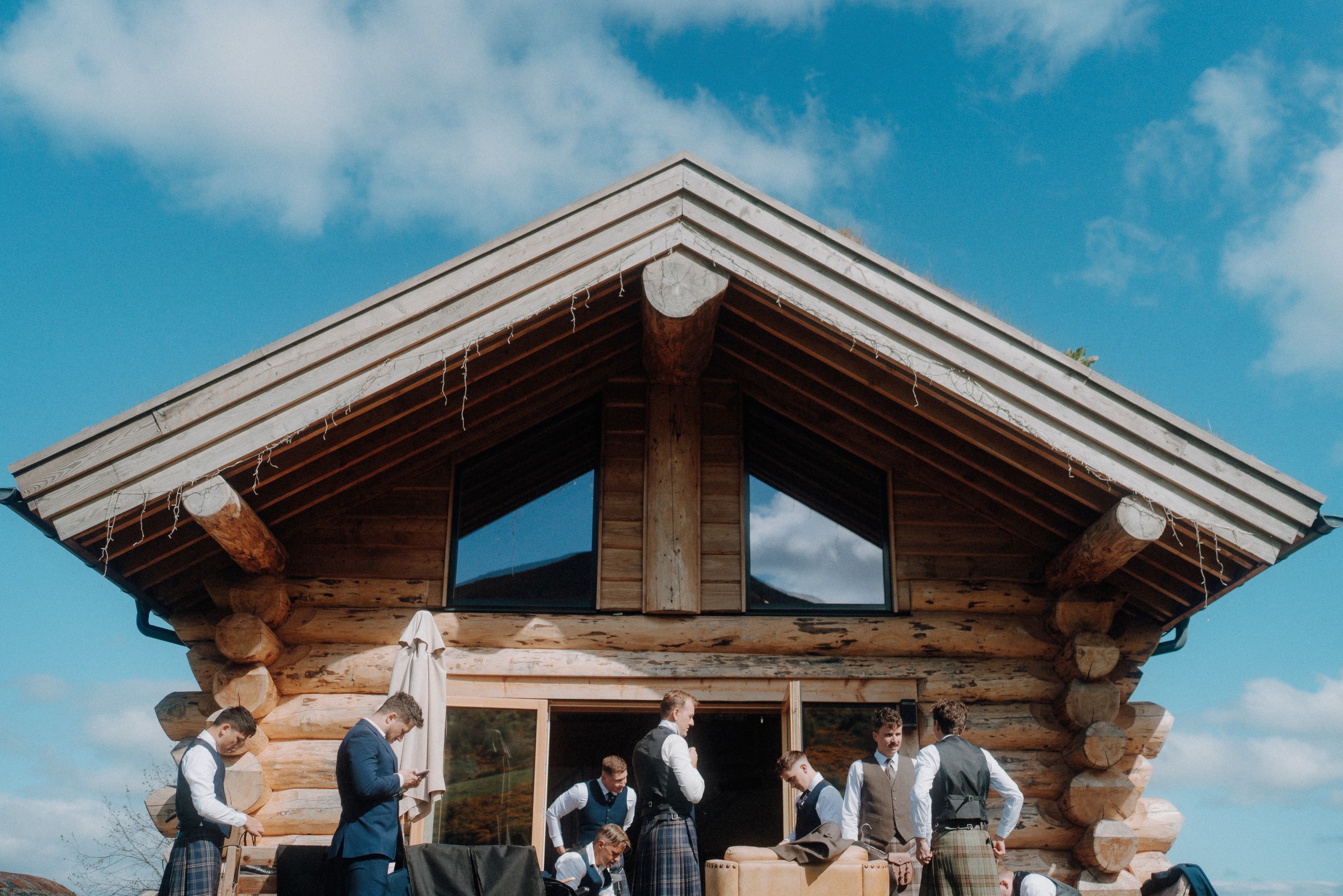 A group of men in formal attire and kilts stand outside a wooden log cabin under a blue sky with scattered clouds.
