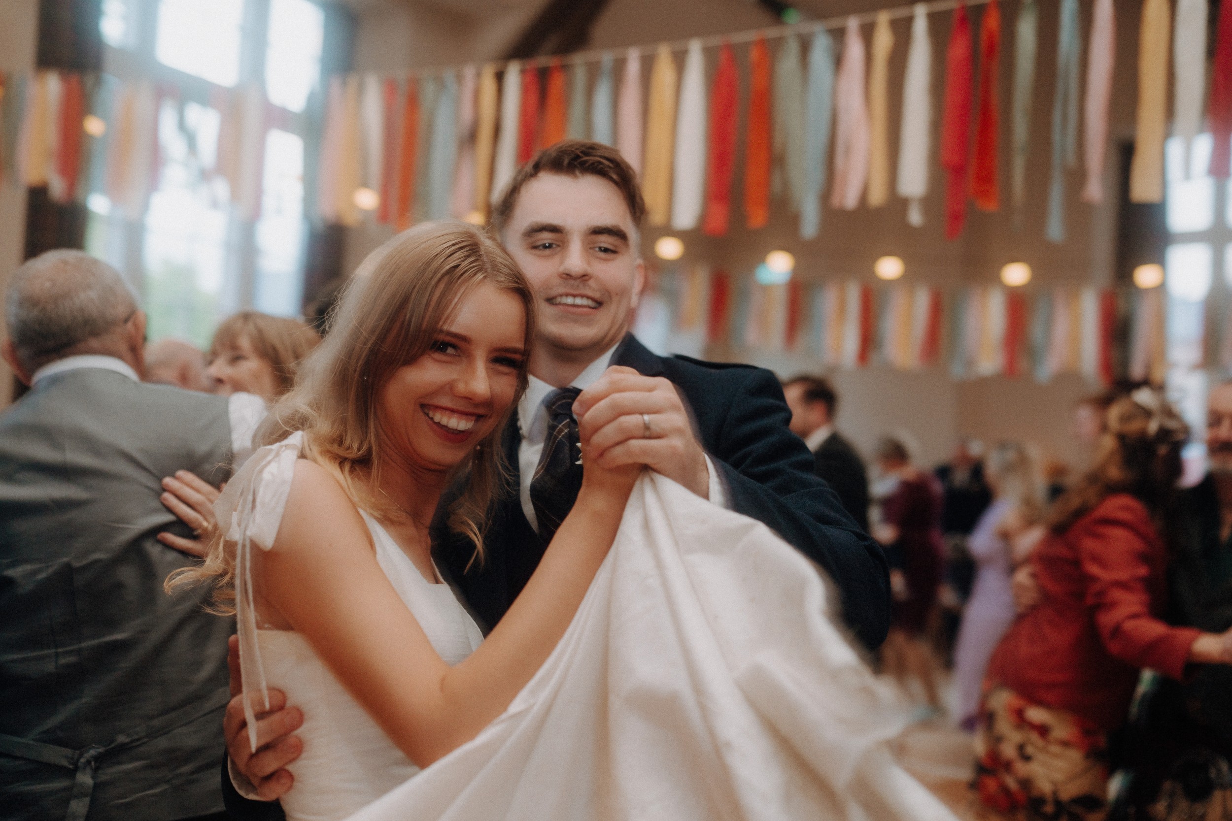 A smiling couple dances together at an indoor event, surrounded by other guests and colorful hanging ribbons in the background.