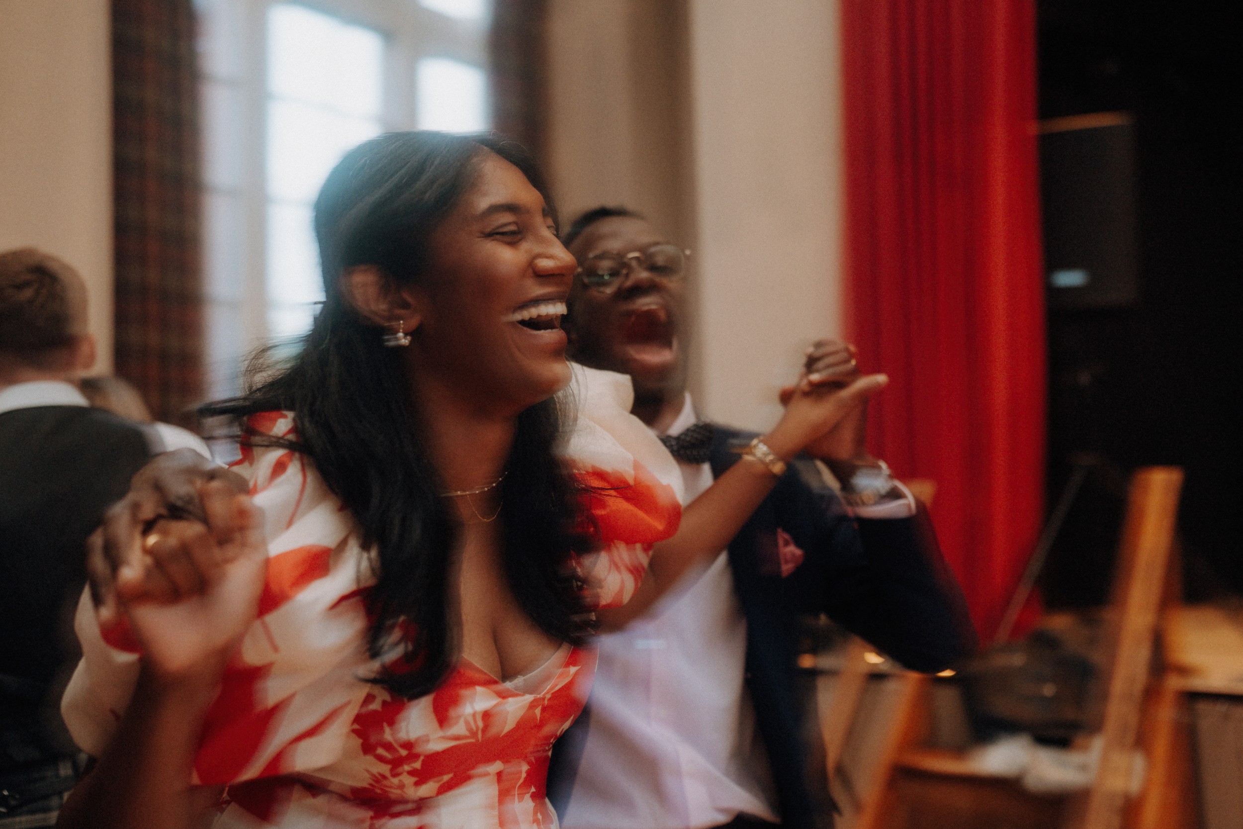 Two people holding hands and laughing at an indoor event, with red curtains and a window in the background.