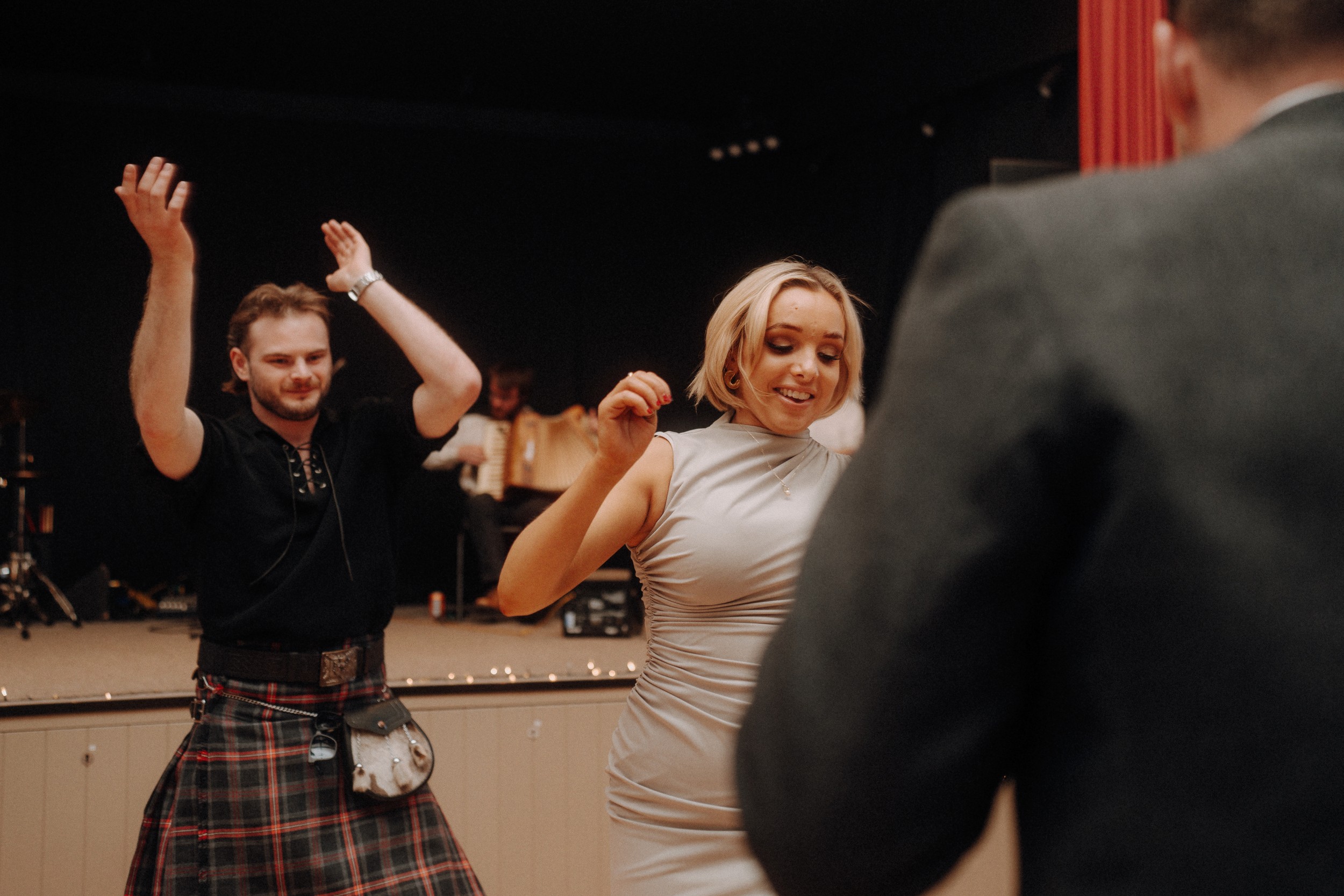 A man in a kilt claps while a woman in a light dress dances indoors, with another person partially visible in the foreground.