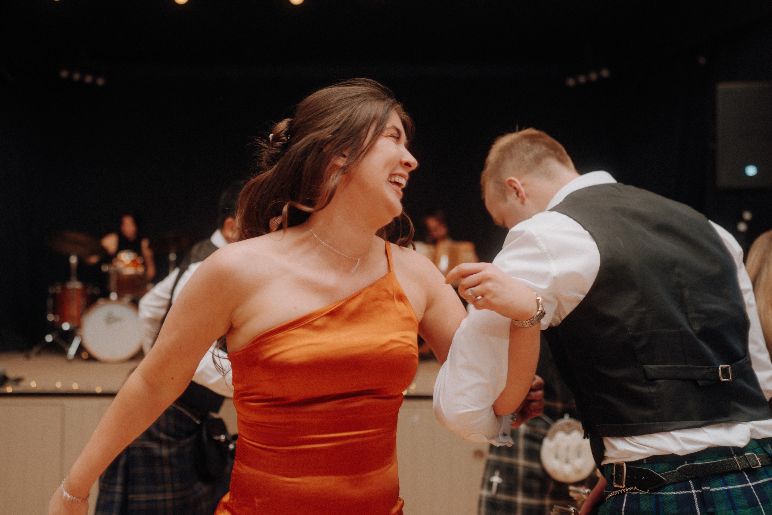 A woman in an orange dress and a man in a vest dance together at an indoor event, with a band playing in the background.