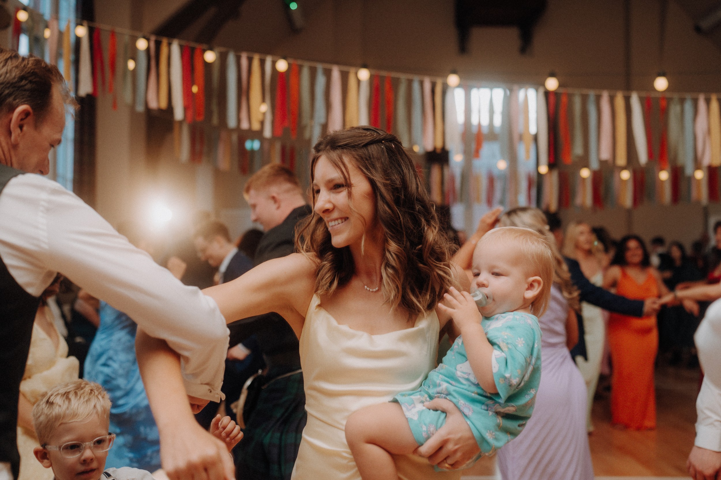 A woman in a white dress holds a small child while dancing at an indoor event with colorful ribbon decorations and other people in the background.