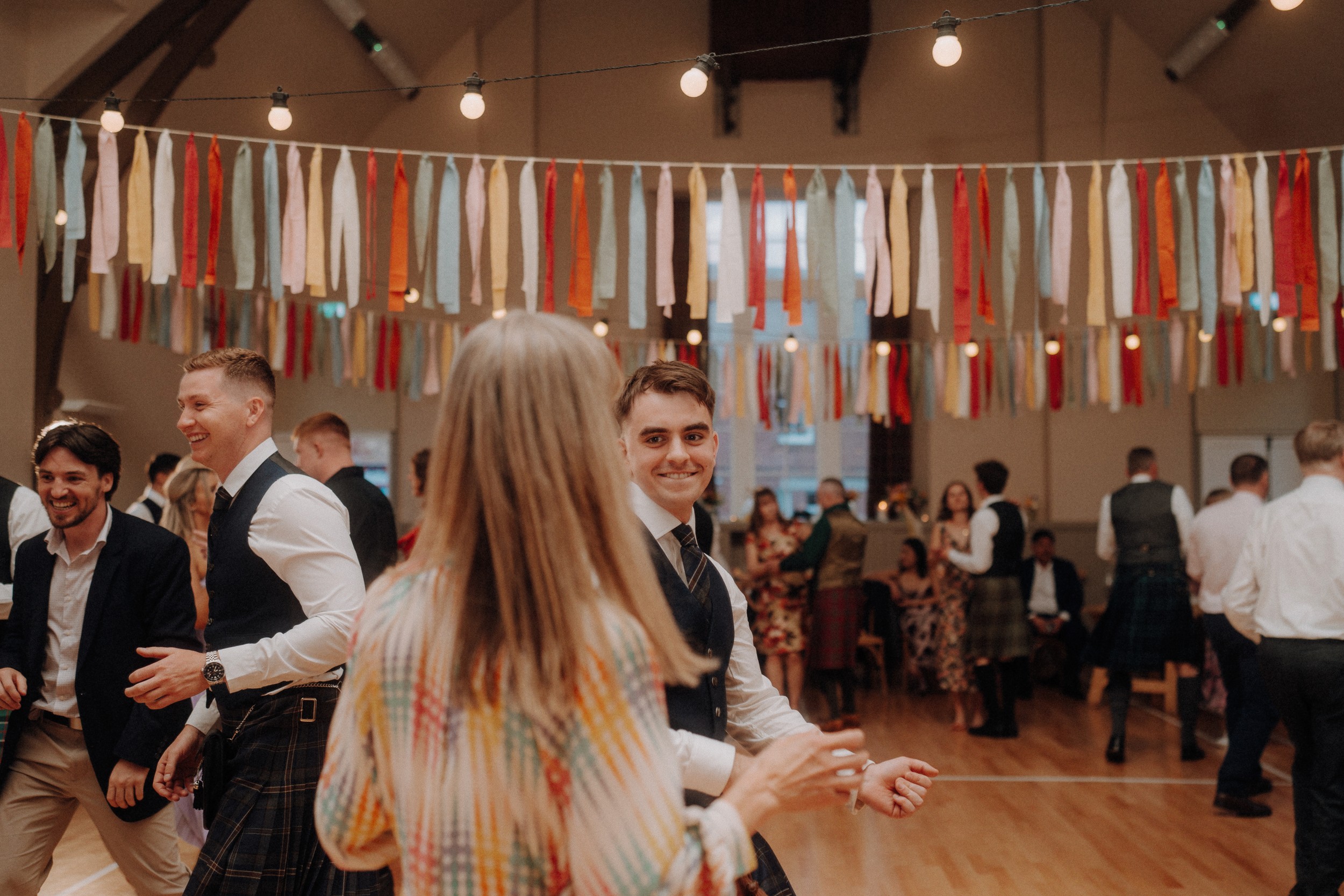 People dance in a decorated hall with colorful hanging ribbons. Most are dressed in formal or semi-formal attire, and the atmosphere appears lively and social.