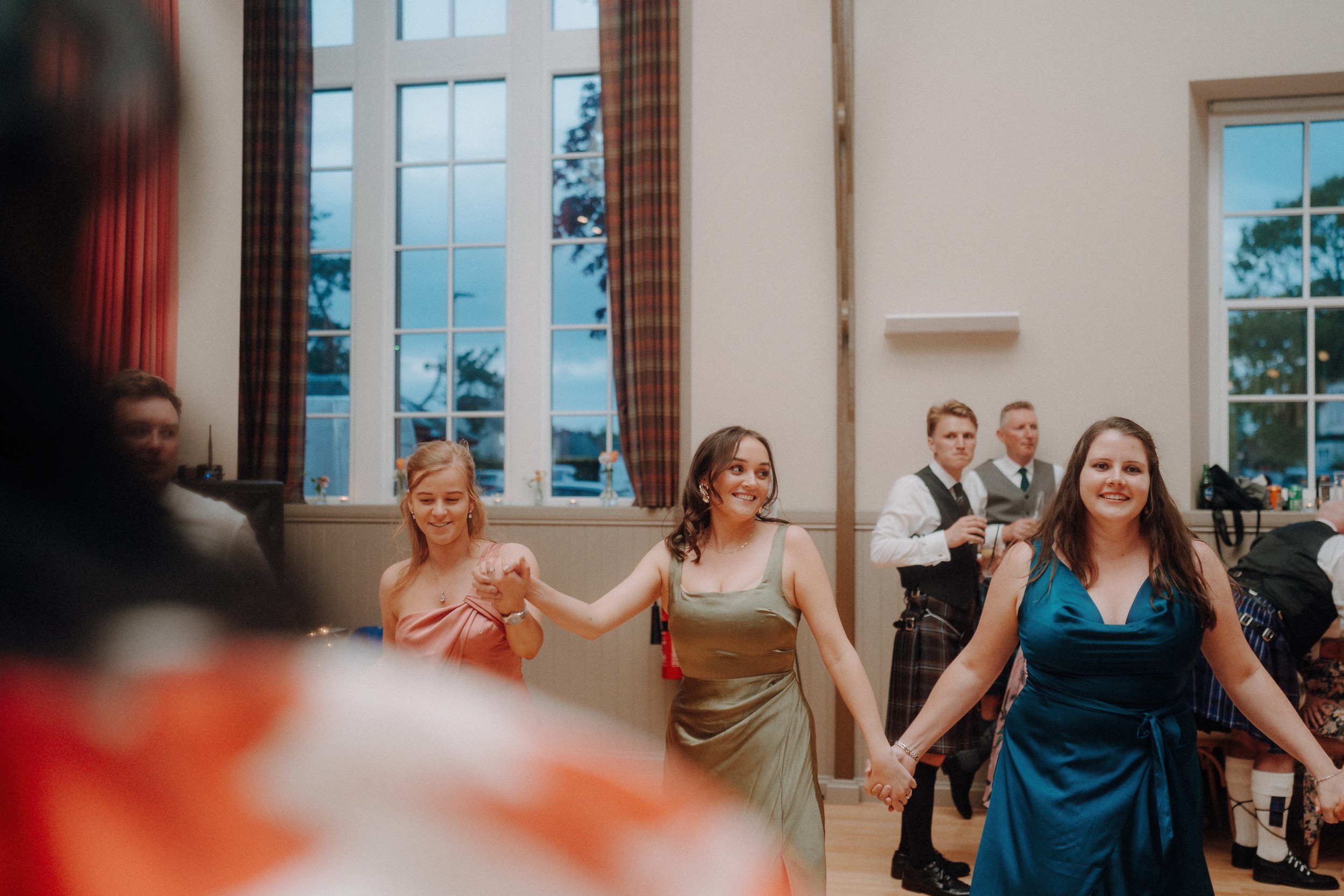 Three women in colorful dresses hold hands and smile while dancing indoors at an event; several men in kilts stand in the background.