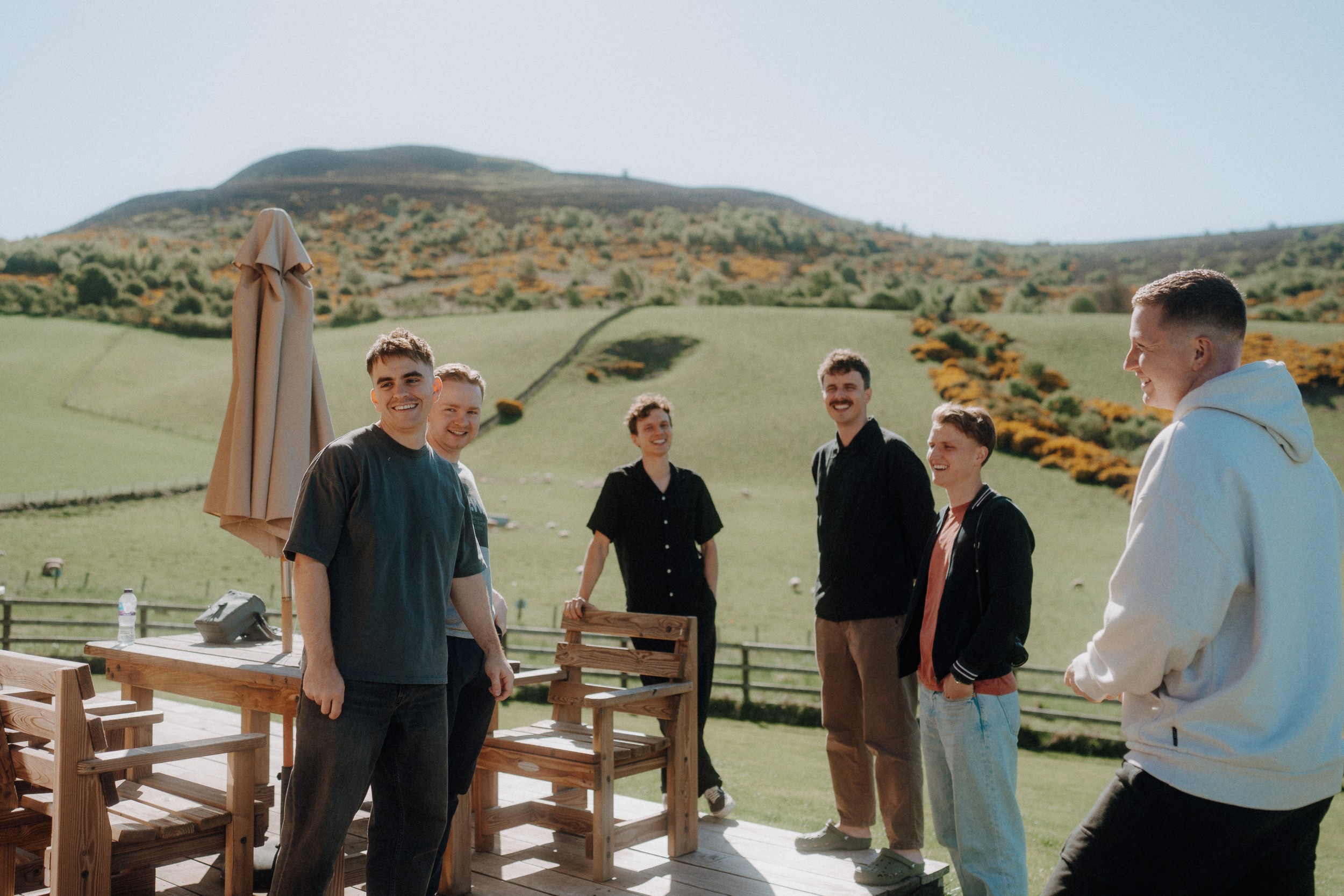 Six people stand and smile on a wooden deck with outdoor furniture, set against a backdrop of green hills and a clear sky.