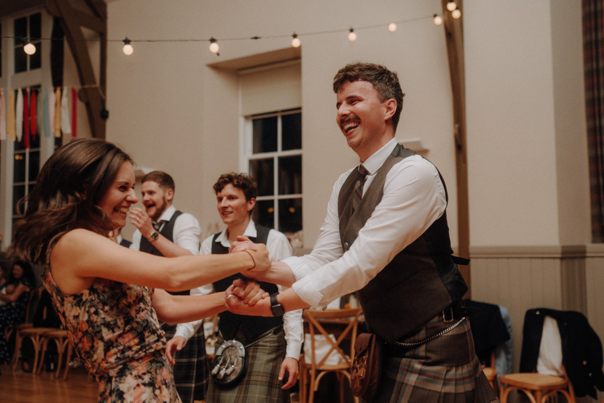 A woman in a floral dress and a man in a kilt dance together in a decorated indoor venue, with others smiling and watching in the background.