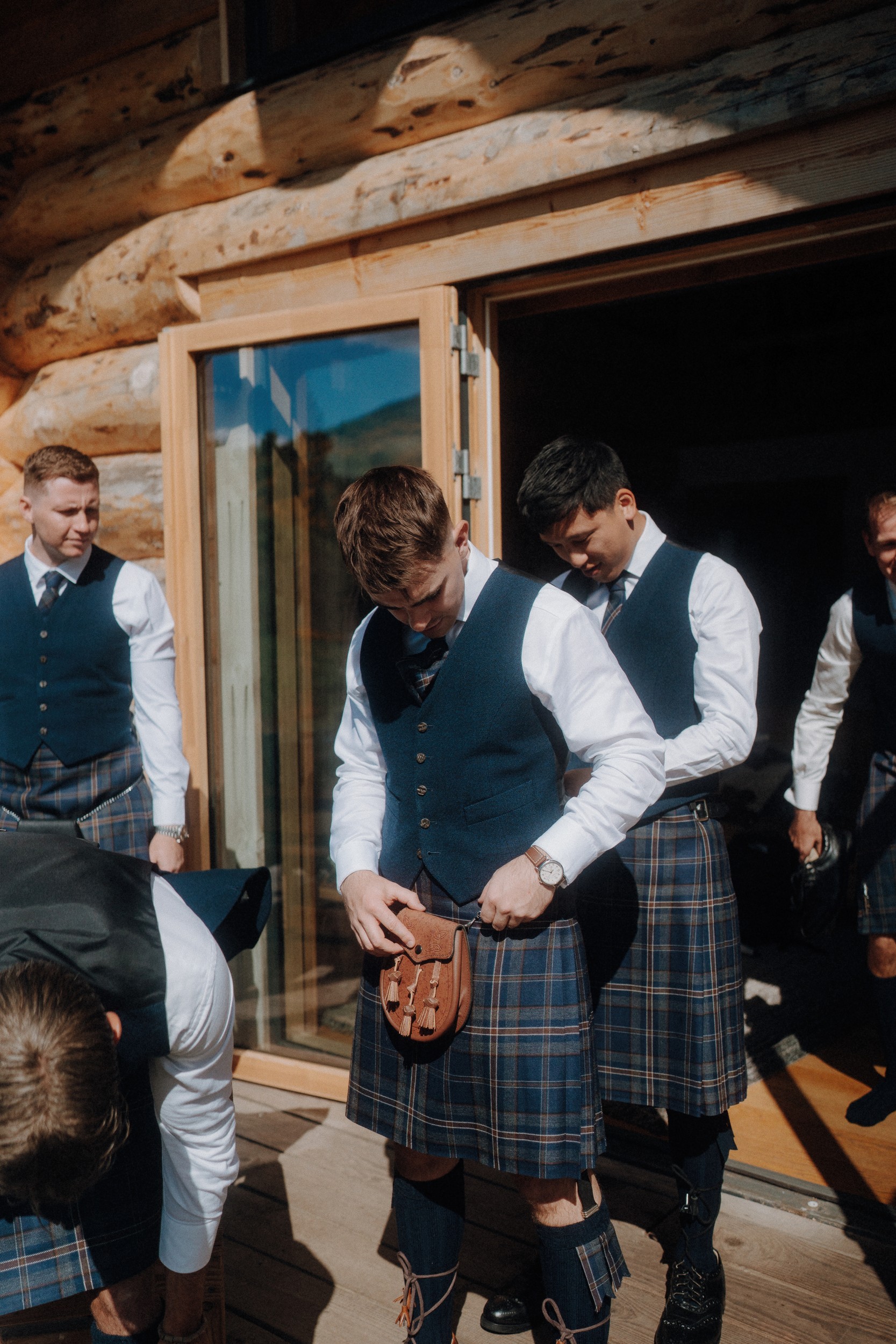 A group of men in matching kilts and vests stand outside a wooden building, adjusting their outfits and accessories in the sunlight.