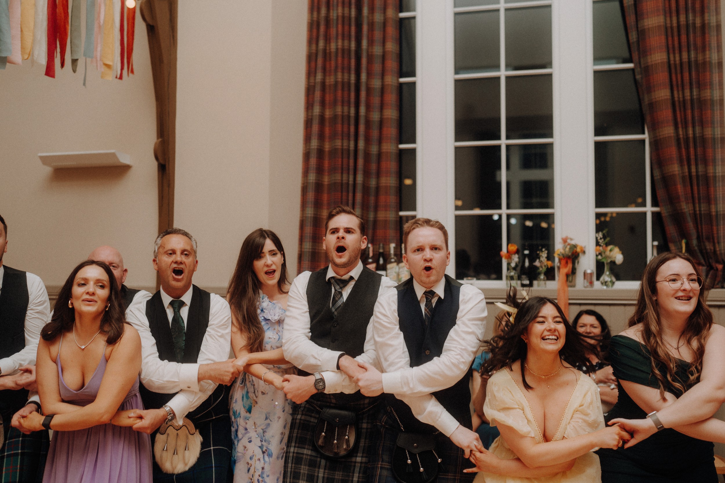 A group of people in formal attire stand in a line with arms linked, singing together at an indoor event with large windows and plaid curtains.