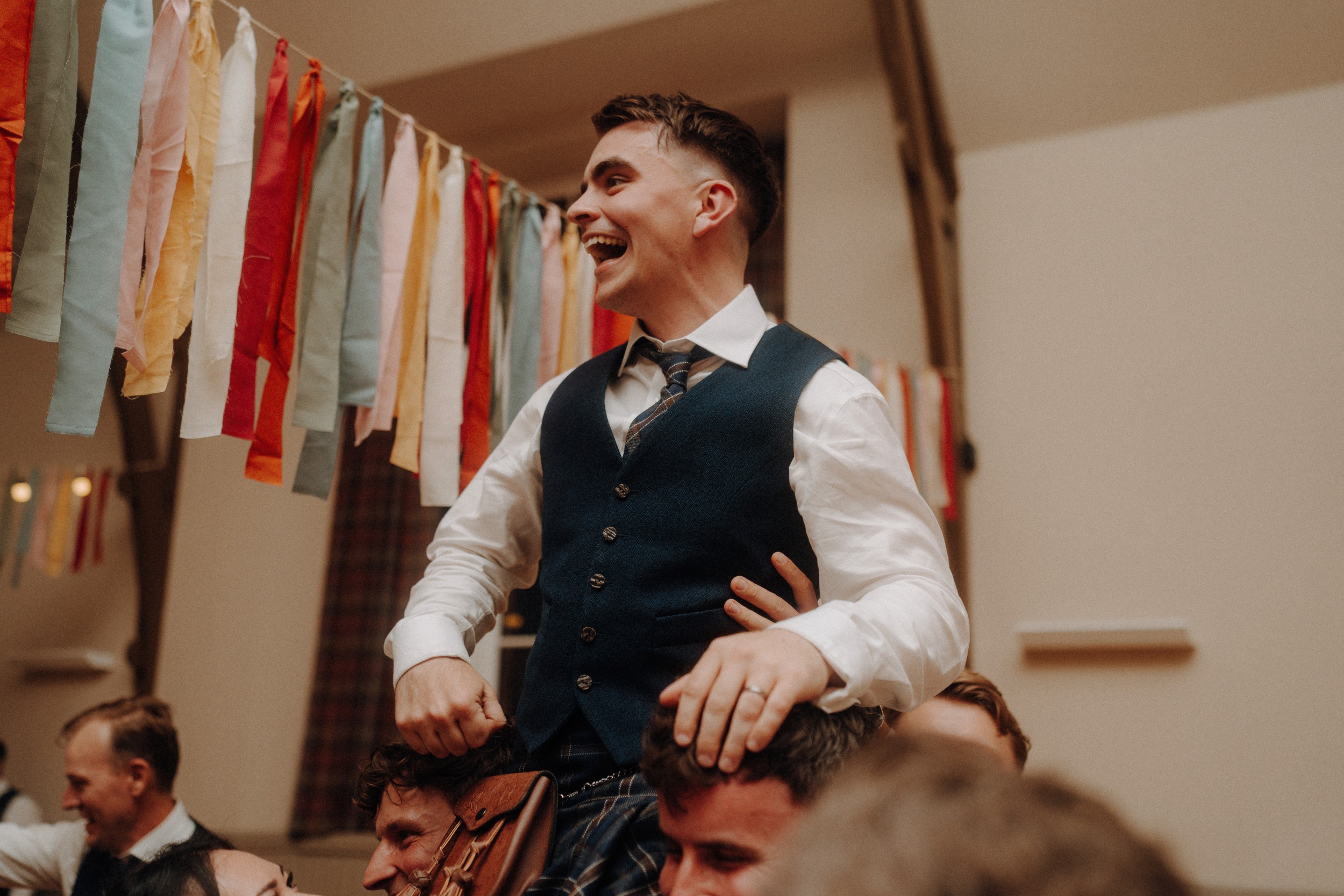 A smiling man in a vest and tie is lifted on the shoulders of others at an indoor celebration decorated with colorful hanging ribbons.