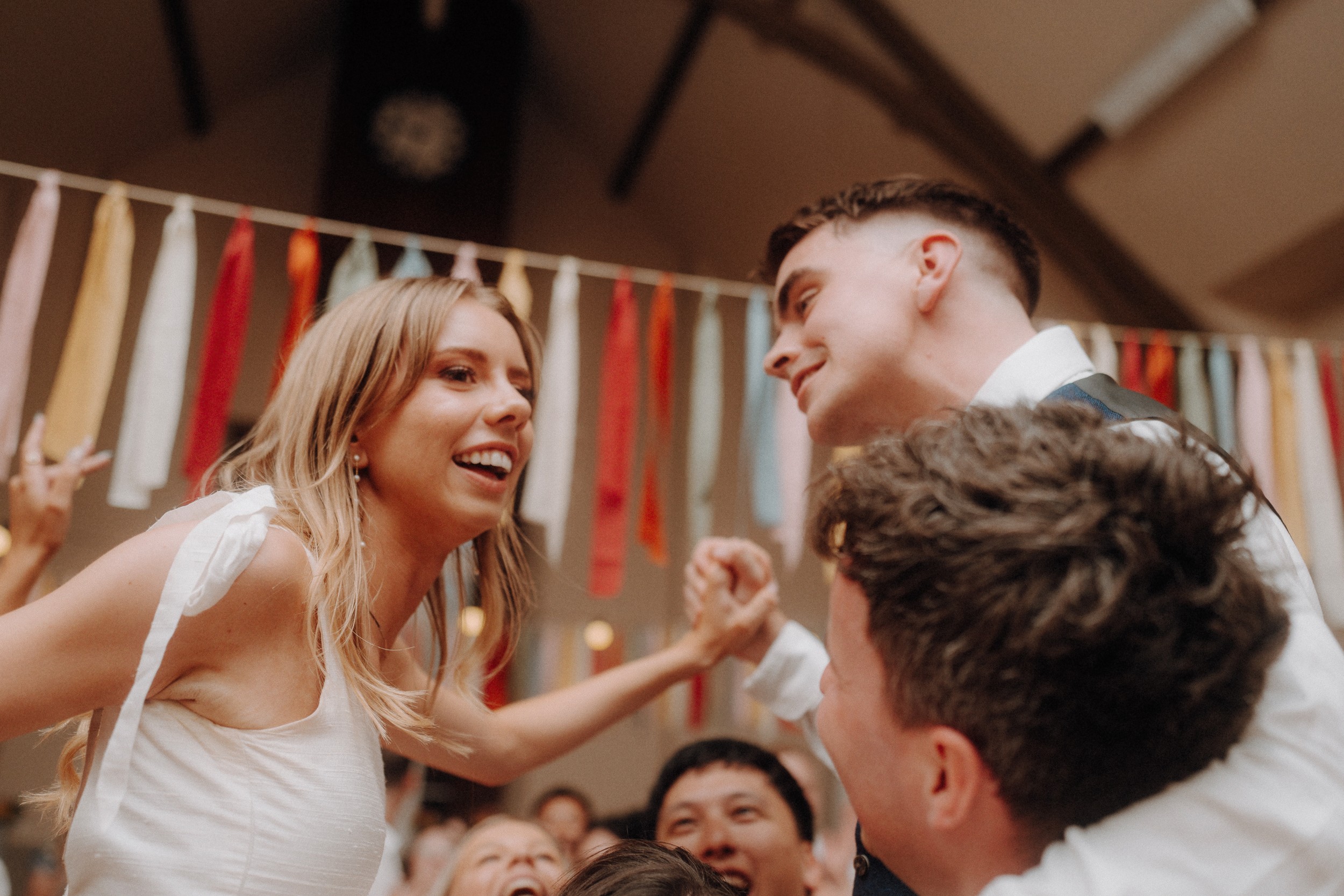 A woman in a white dress and a man in a suit are lifted on chairs at a lively celebration, surrounded by people and colorful hanging ribbons in the background.