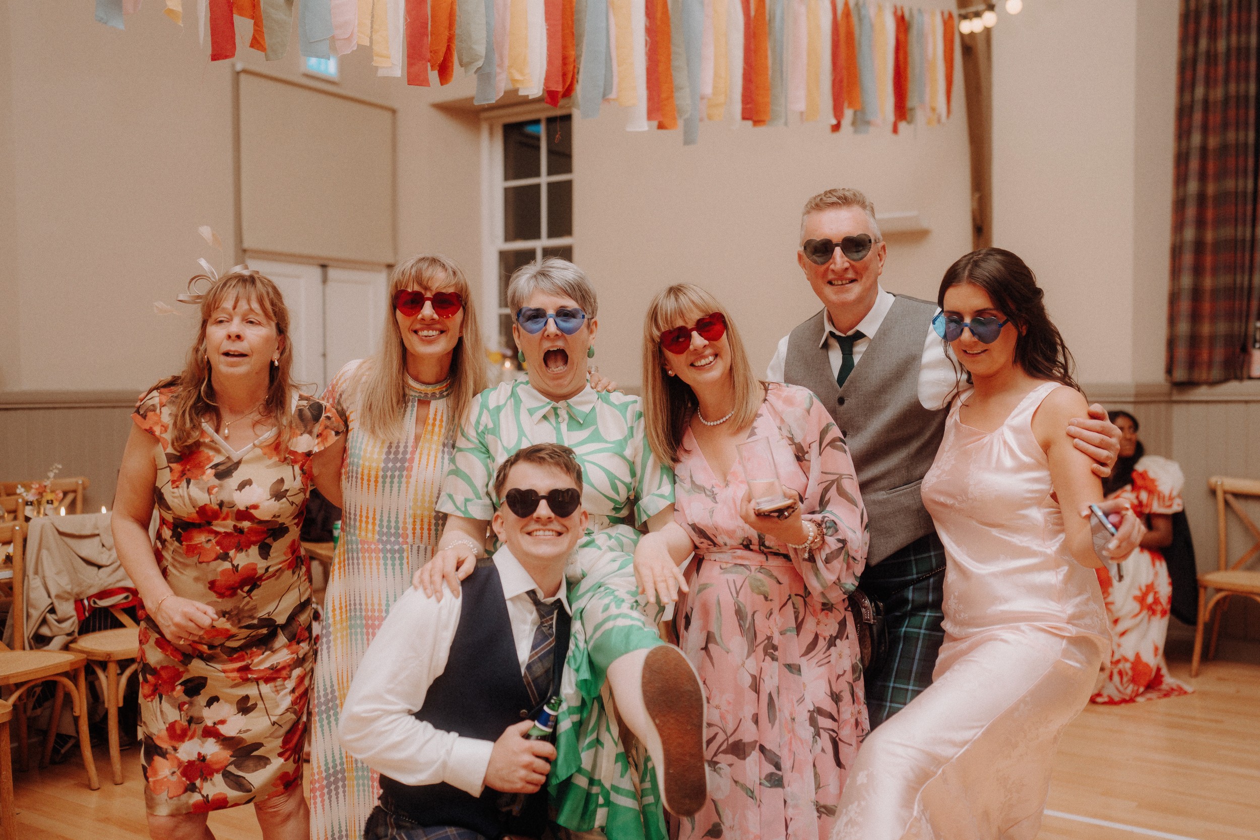 Seven people in colorful outfits and heart-shaped sunglasses pose together and smile in a decorated indoor venue with streamers hanging from the ceiling.