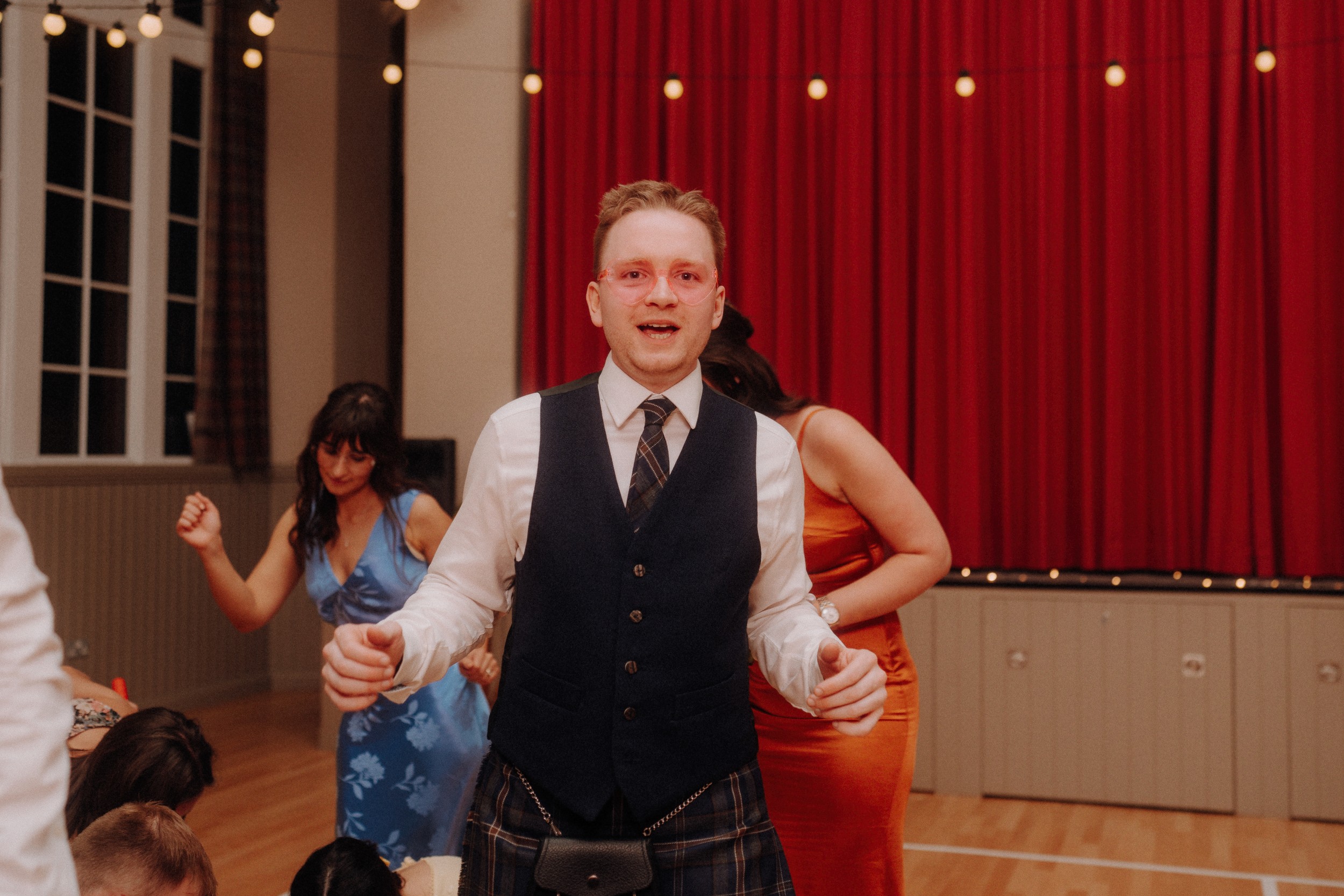 A man in a vest, tie, and kilt stands indoors with a red curtain behind him. People in colorful dresses are dancing in the background. String lights hang overhead.