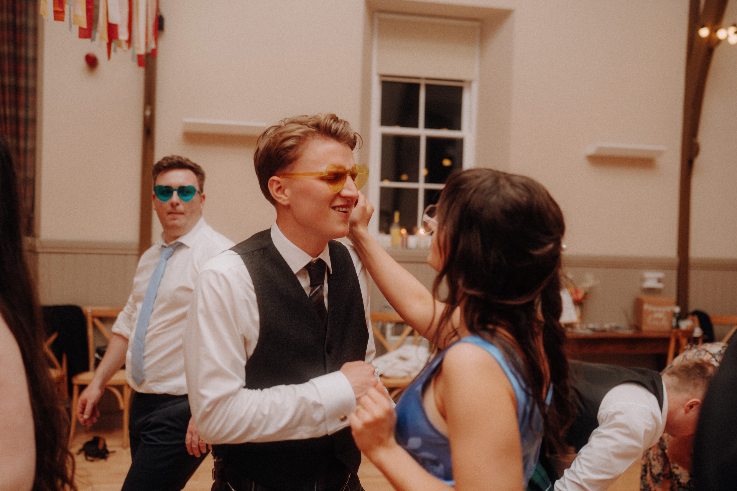 Three people wearing sunglasses and formal attire are dancing indoors at an event, with one woman touching a man's face while others are nearby.