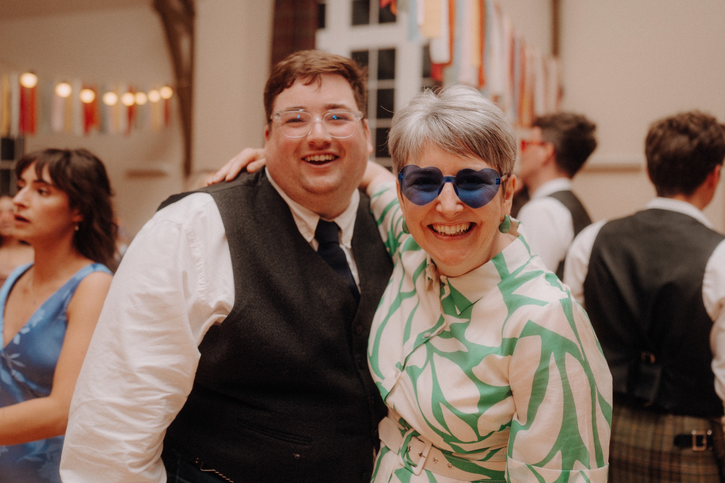 Two people smiling at a social event, one wearing a dark vest and tie, the other in a green and white outfit with blue sunglasses. Other guests are in the background.