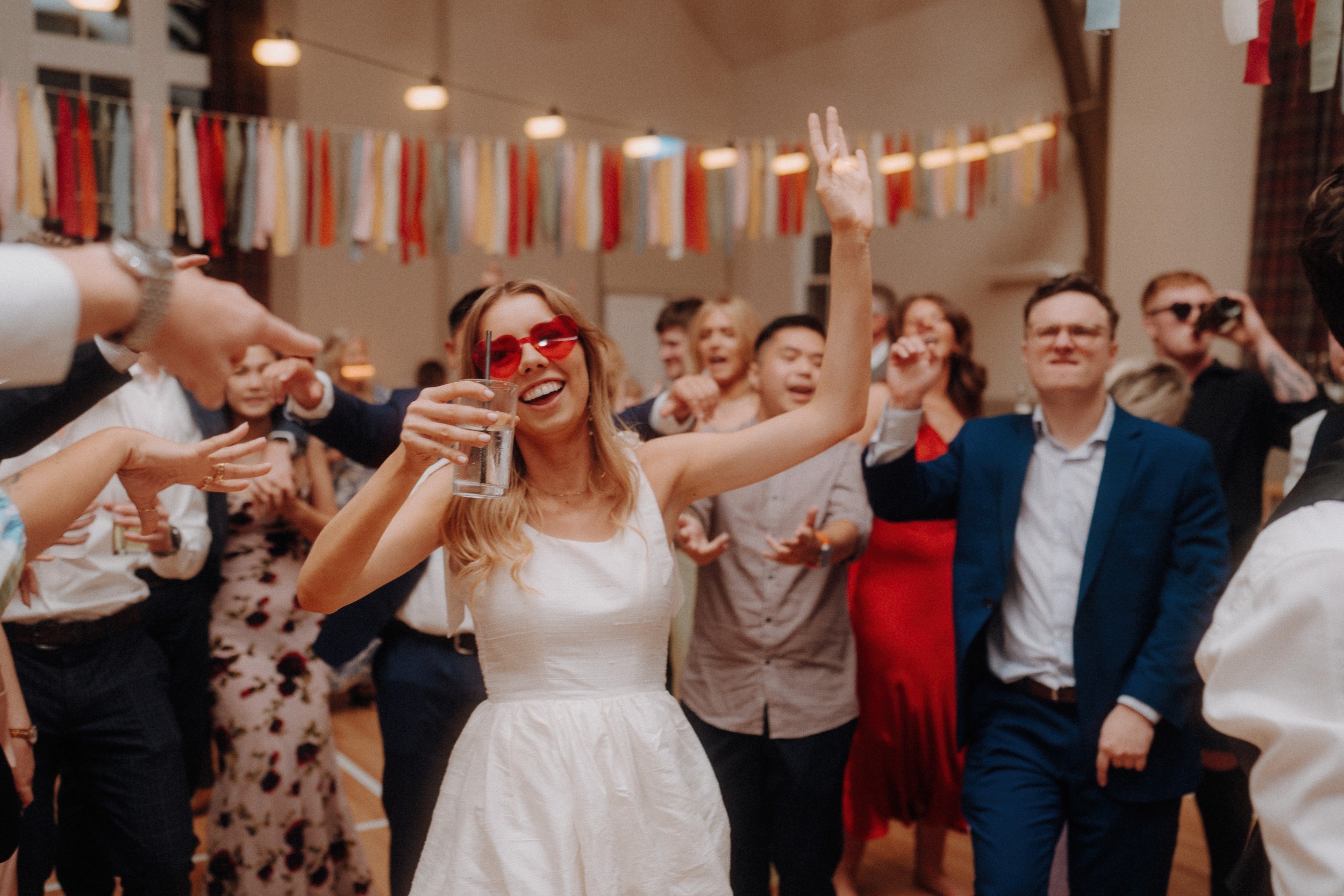 A woman in a white dress and red heart-shaped sunglasses holds a drink and dances at a lively indoor party with people in formal attire.