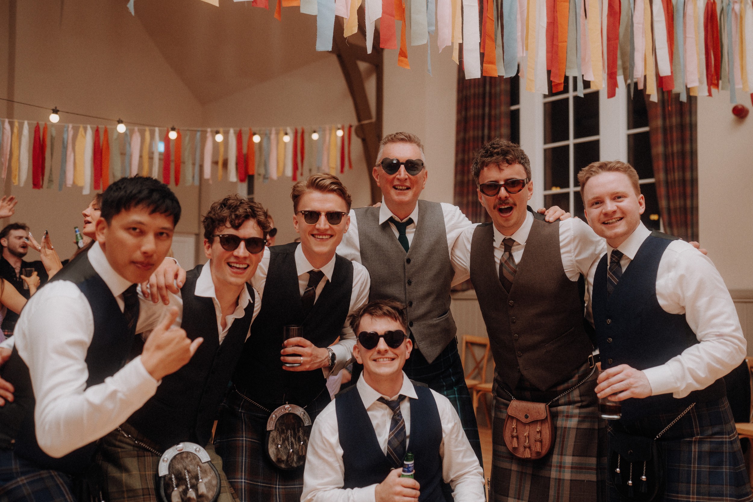 Seven men in kilts and vests pose together at an indoor event, several wearing sunglasses; colorful streamers hang above them.