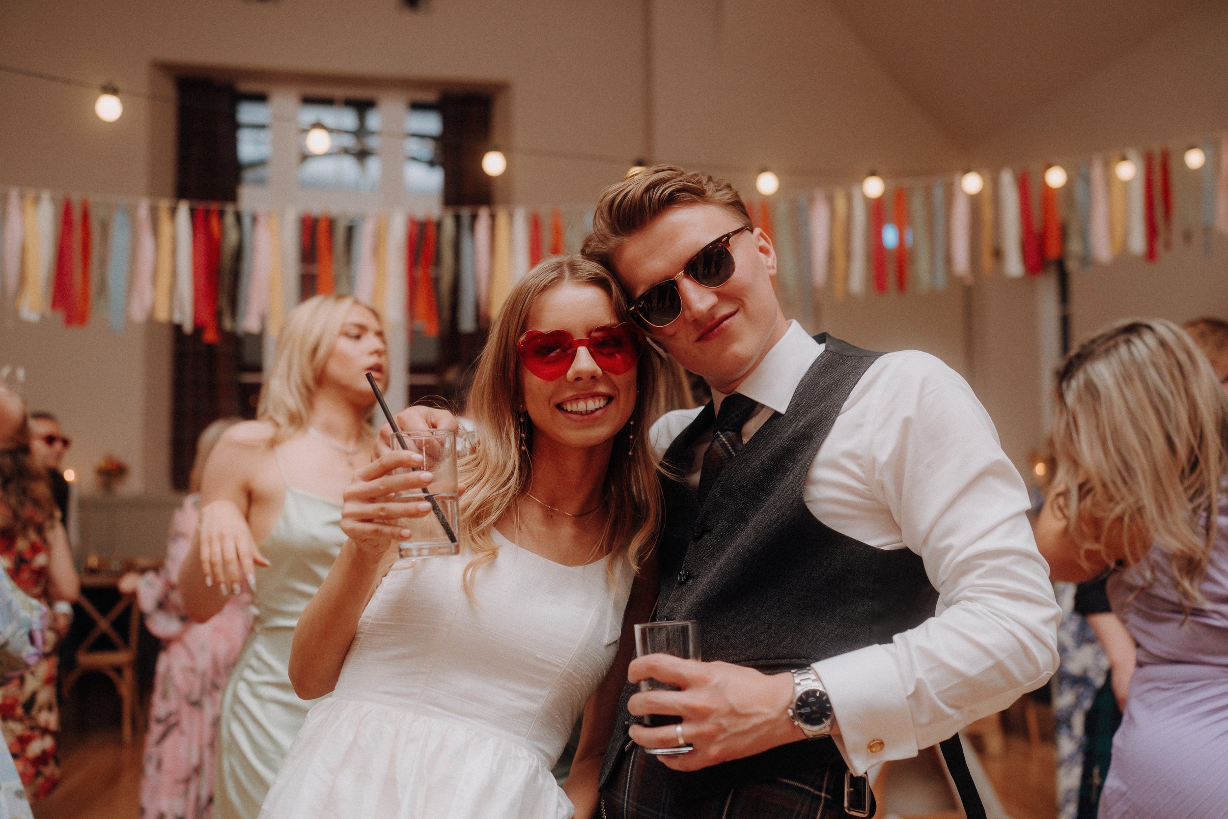 A smiling couple wearing sunglasses poses together at a party, each holding a drink, with colorful streamers and other guests in the background.