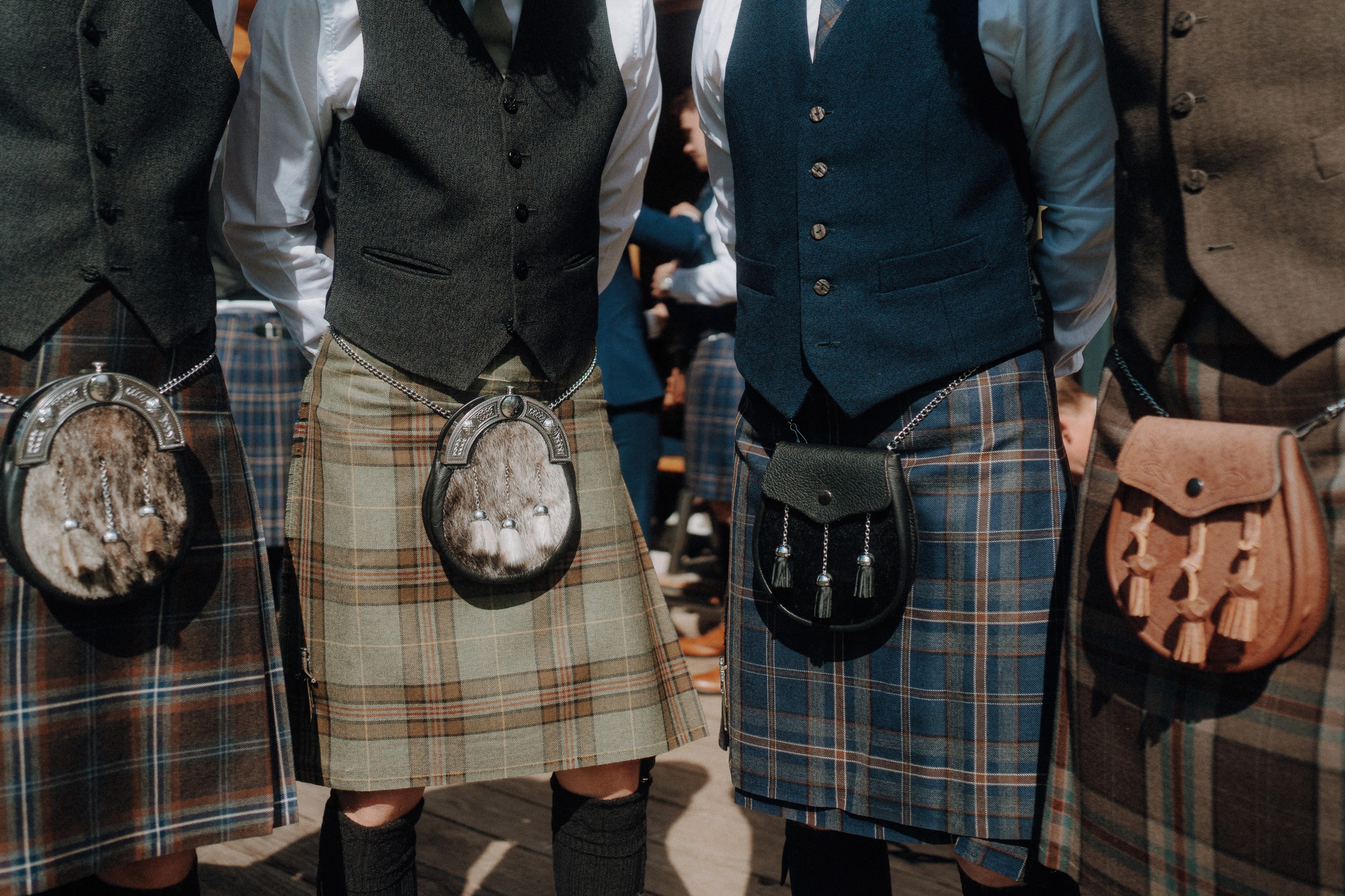 Three people wearing traditional Scottish kilts, waistcoats, and sporrans, standing side by side. Only their torsos and kilts are visible.
