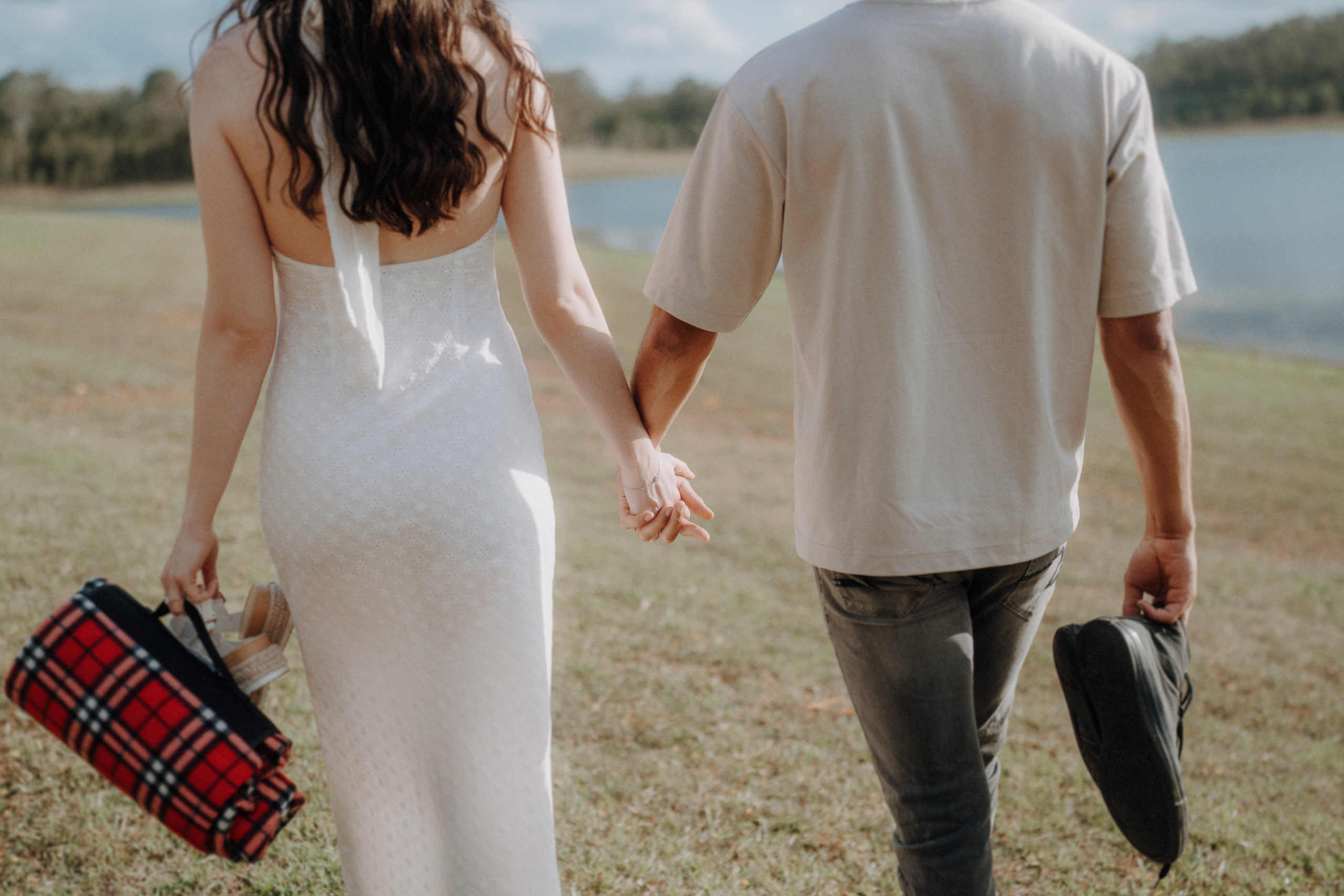 A woman in a white dress and a man in a light shirt walk hand in hand on grass near water; she holds a picnic basket, he holds shoes.