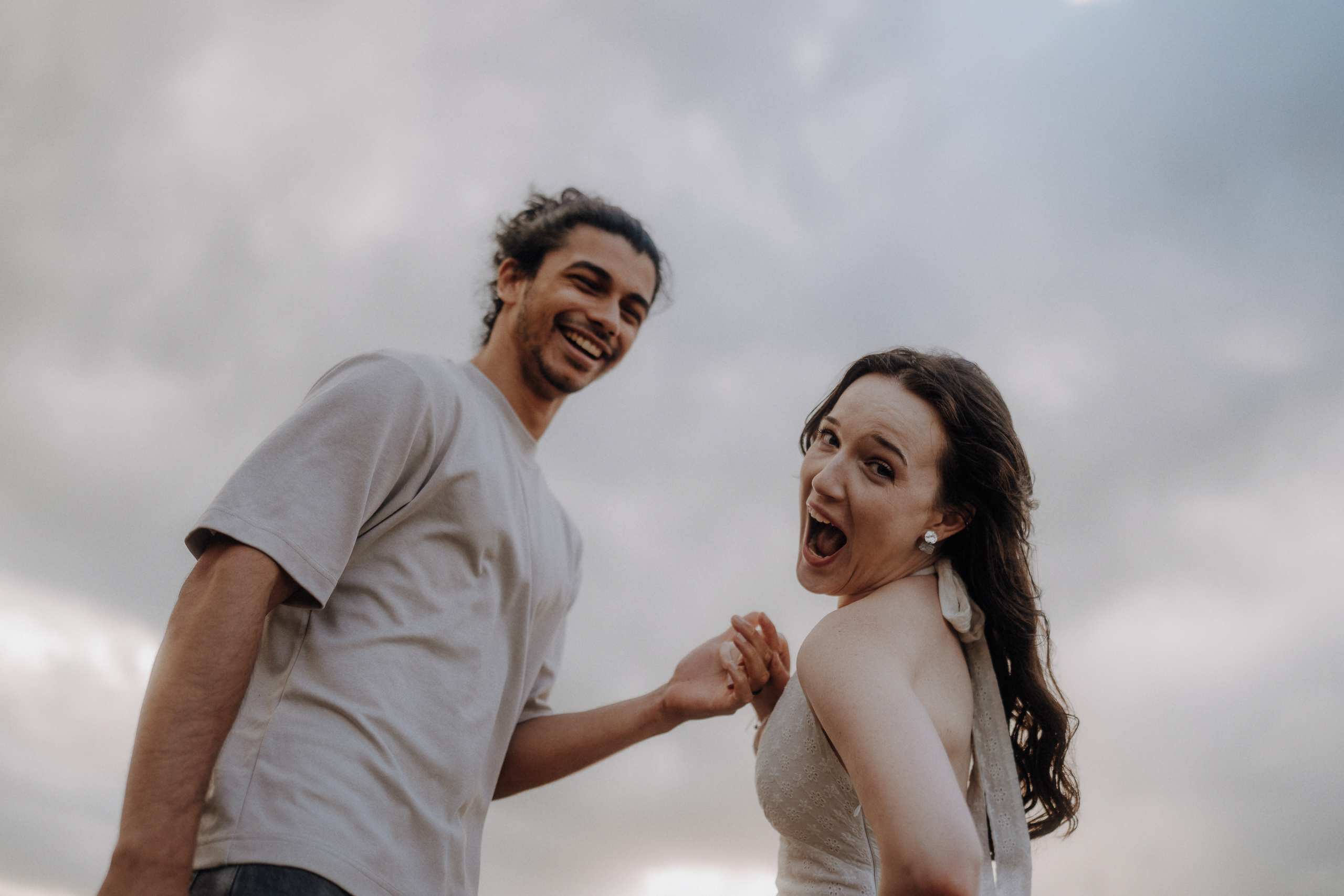 Two people stand outdoors, smiling and laughing, with a cloudy sky in the background.