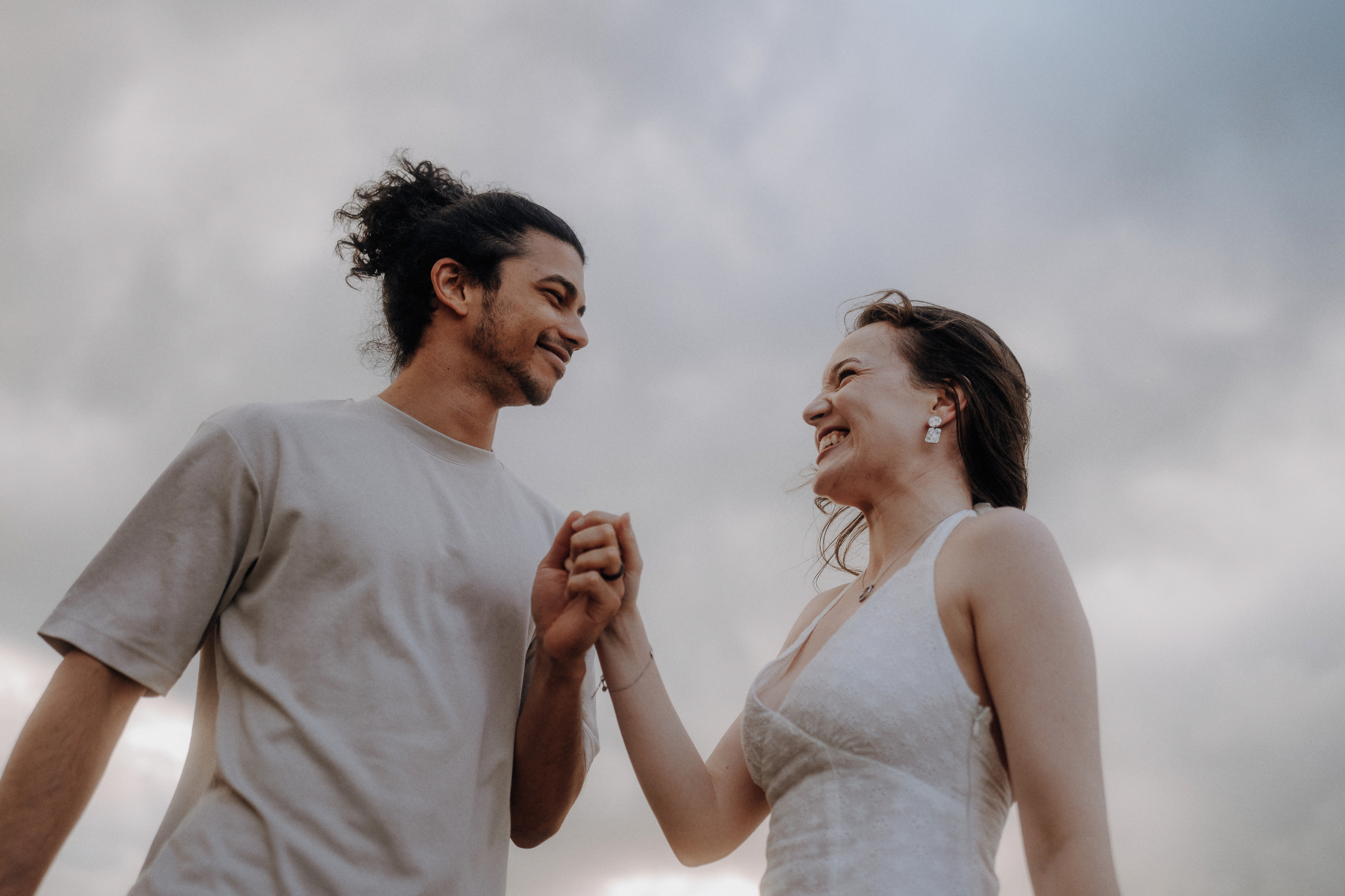 Two people stand outdoors holding hands and smiling at each other, with a cloudy sky in the background.