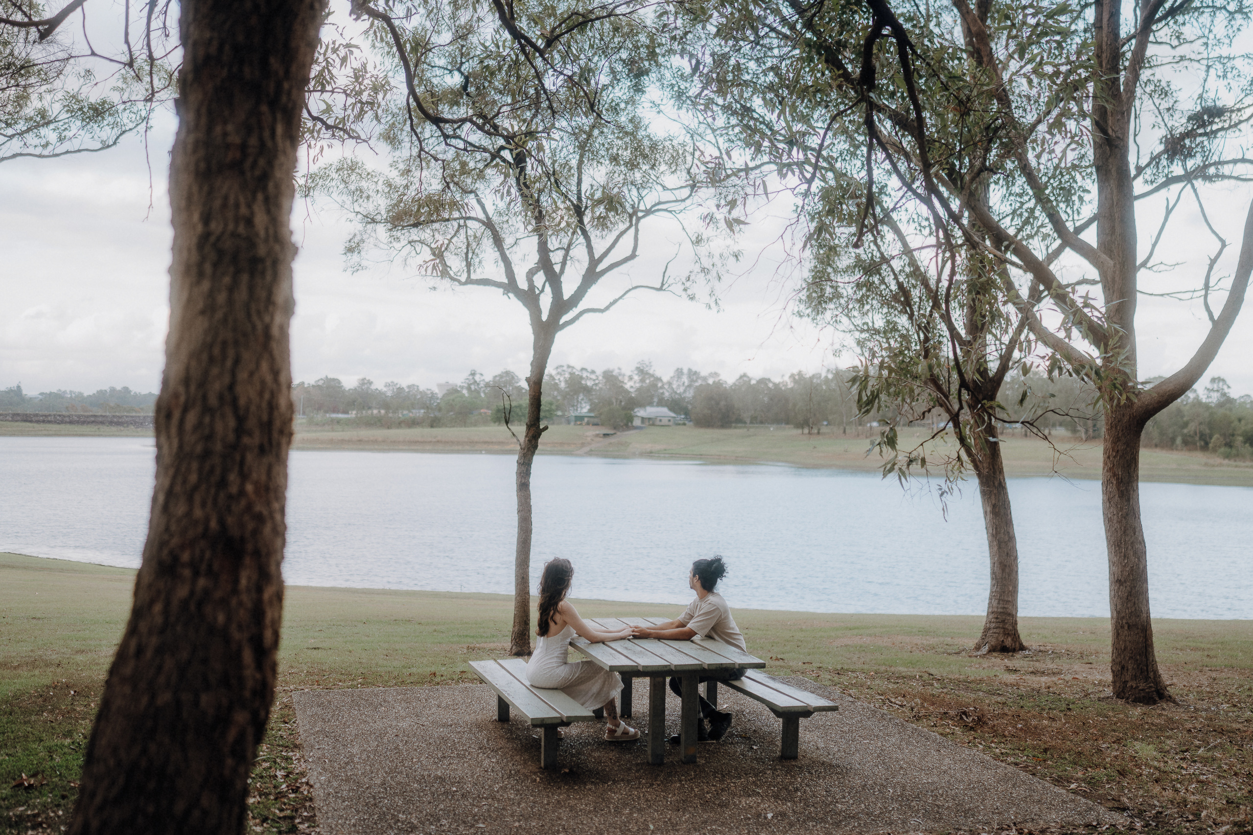Two people sit at a picnic table under trees by a lake, facing each other, with grass and water in the background.