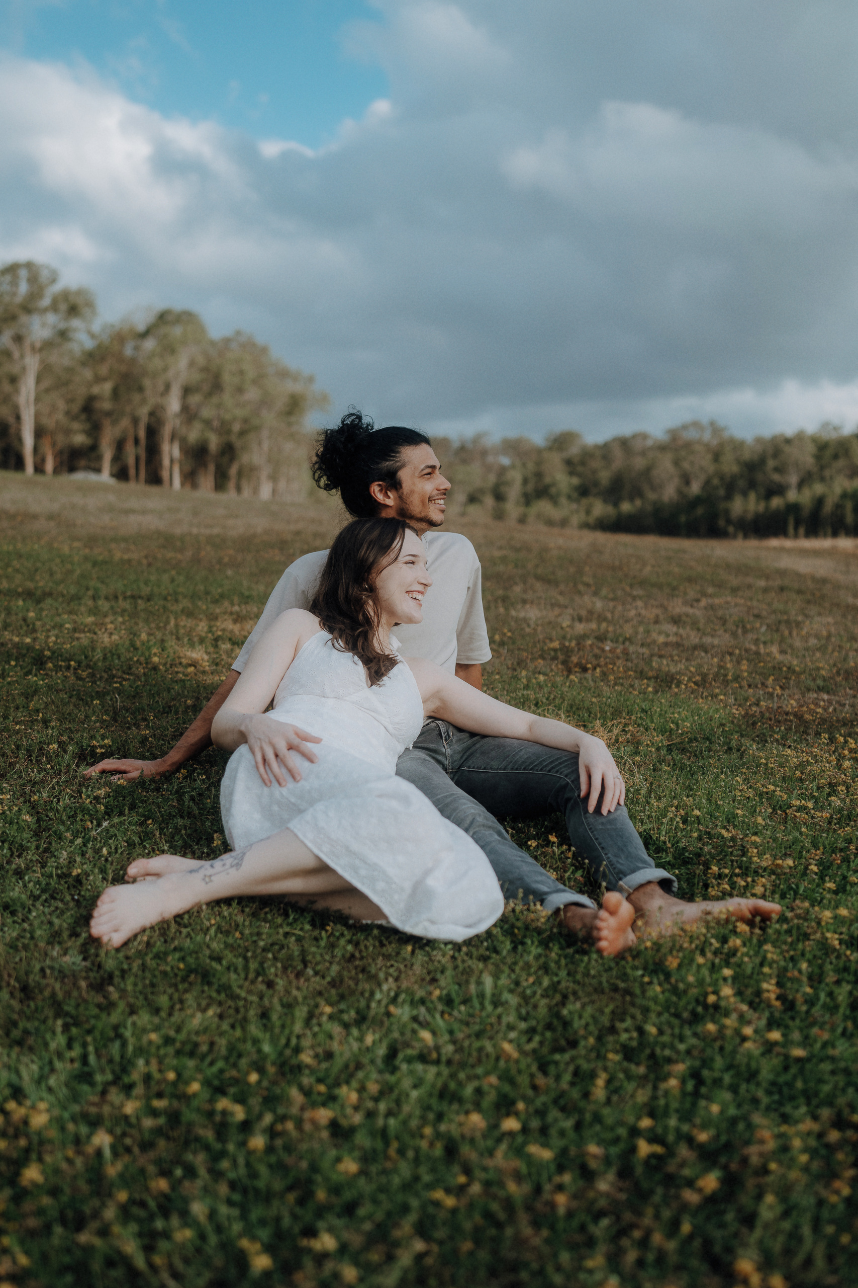 Two people sit barefoot on grass in a field, leaning against each other and looking in the same direction, with trees and clouds in the background.