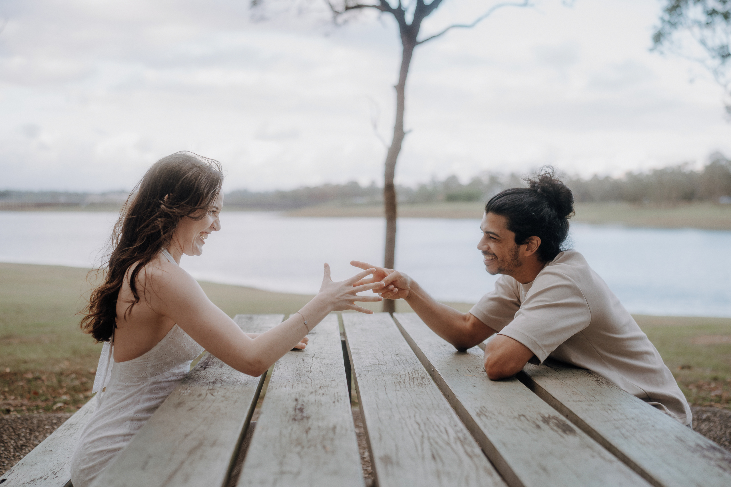 Two people sit across from each other at a wooden picnic table outdoors, smiling and playing a hand game near a lake with trees in the background.