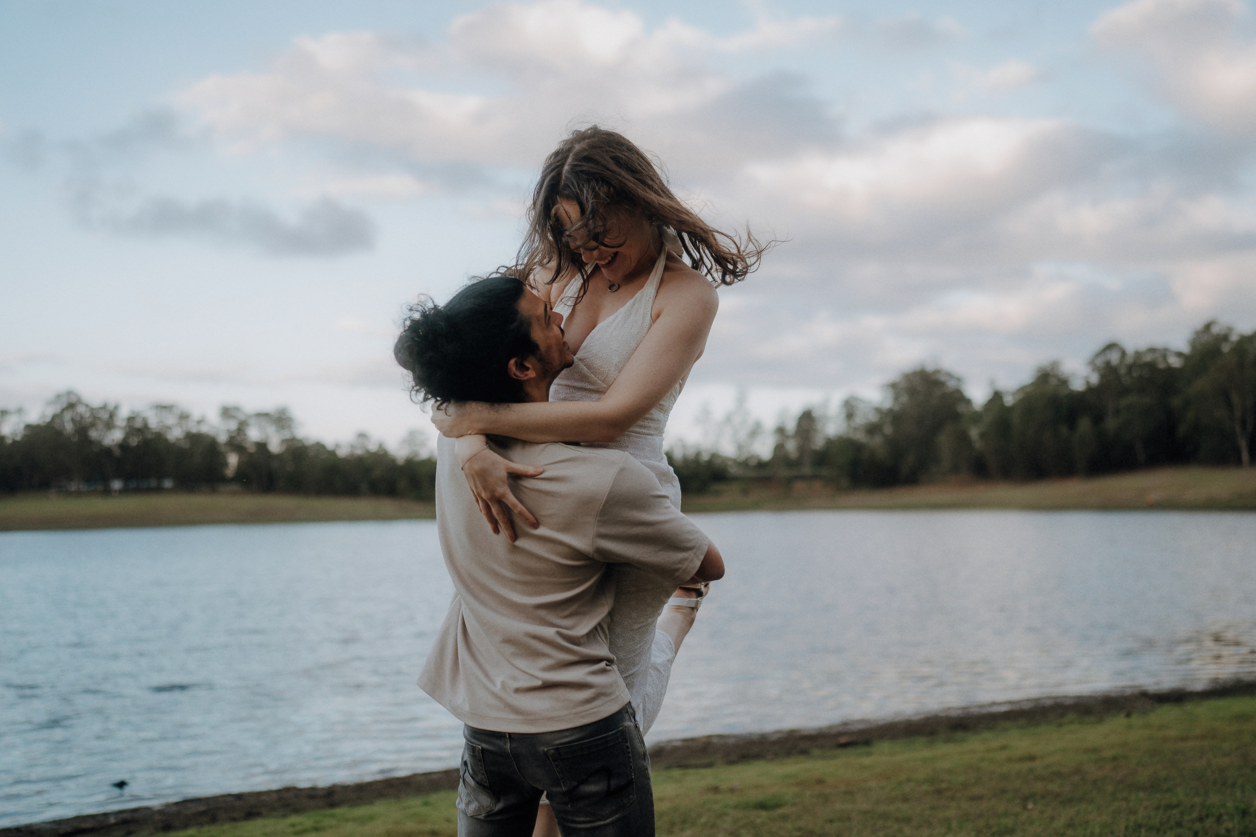 A man lifts a woman in a white dress by a lakeside, both smiling, with trees and water in the background under a cloudy sky.