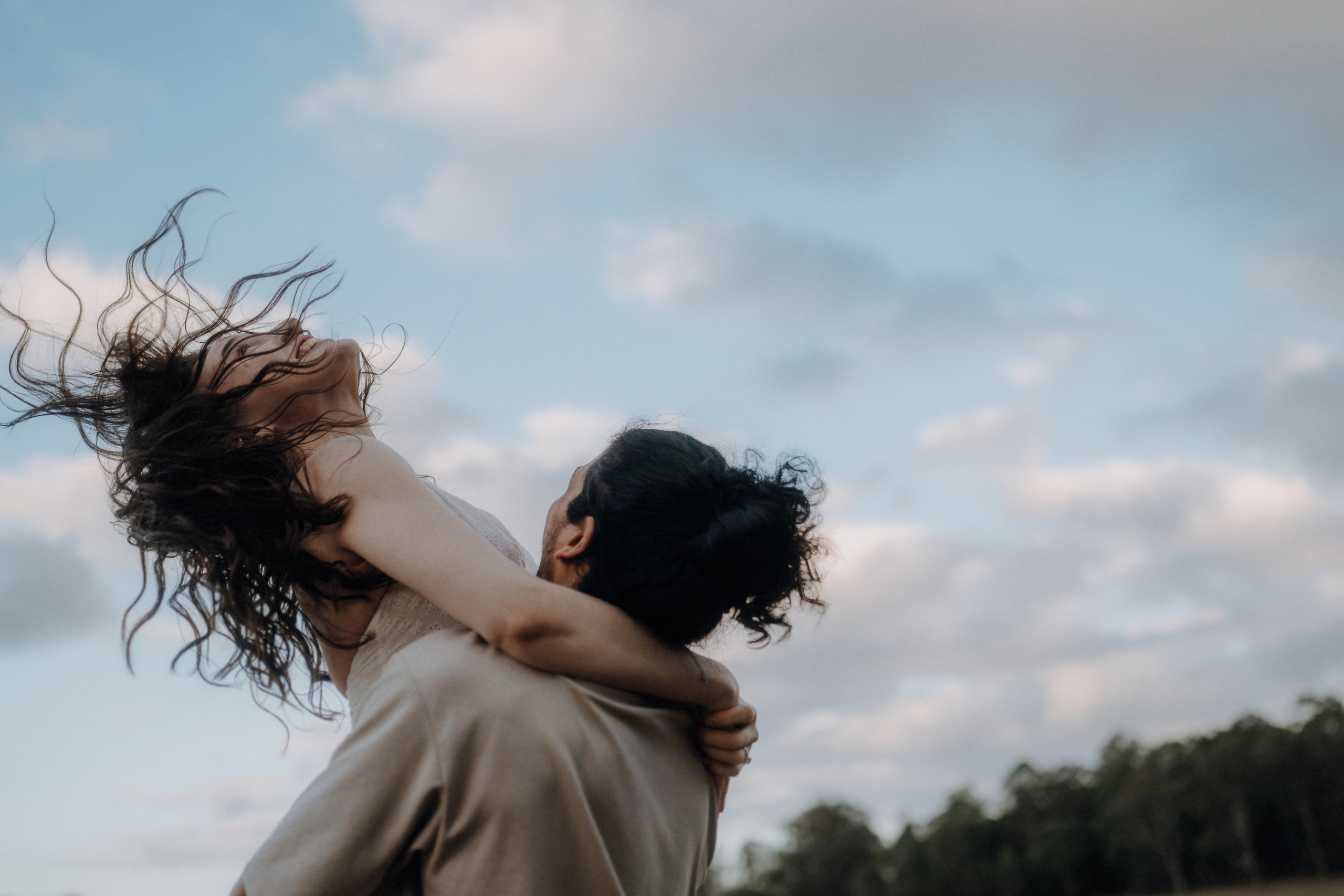 A person lifts another person in their arms outdoors, with both facing away from the camera and a cloudy sky in the background.