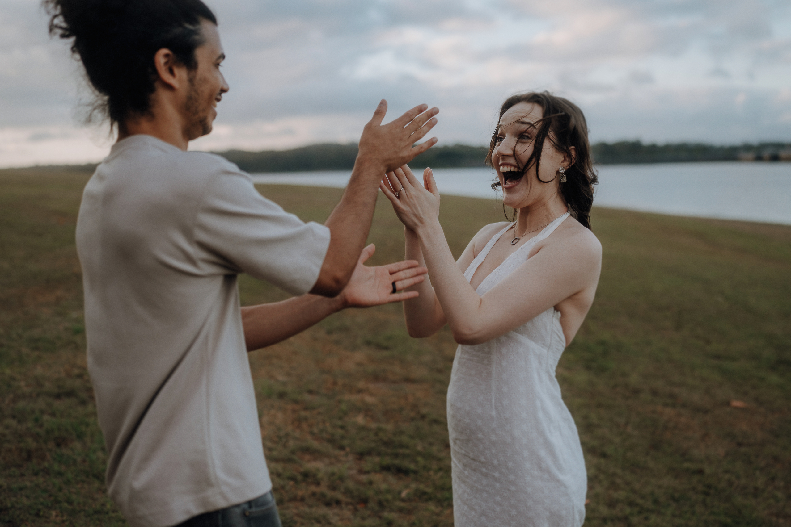 A man and a woman stand on grass near a body of water, facing each other and laughing while playing a hand-clapping game.