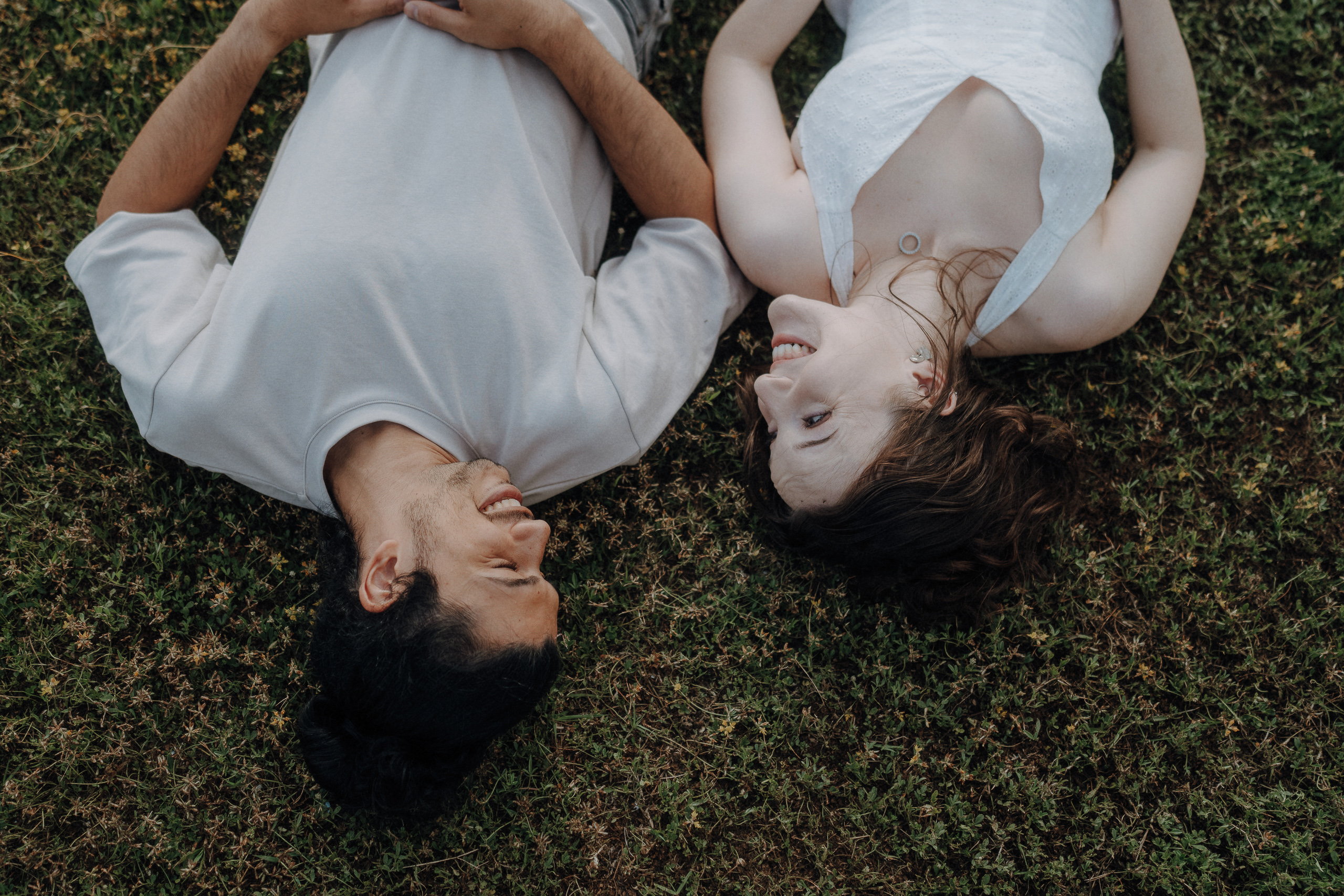 Two people lie on grass facing each other, smiling and making eye contact, with their hands resting on their stomachs.