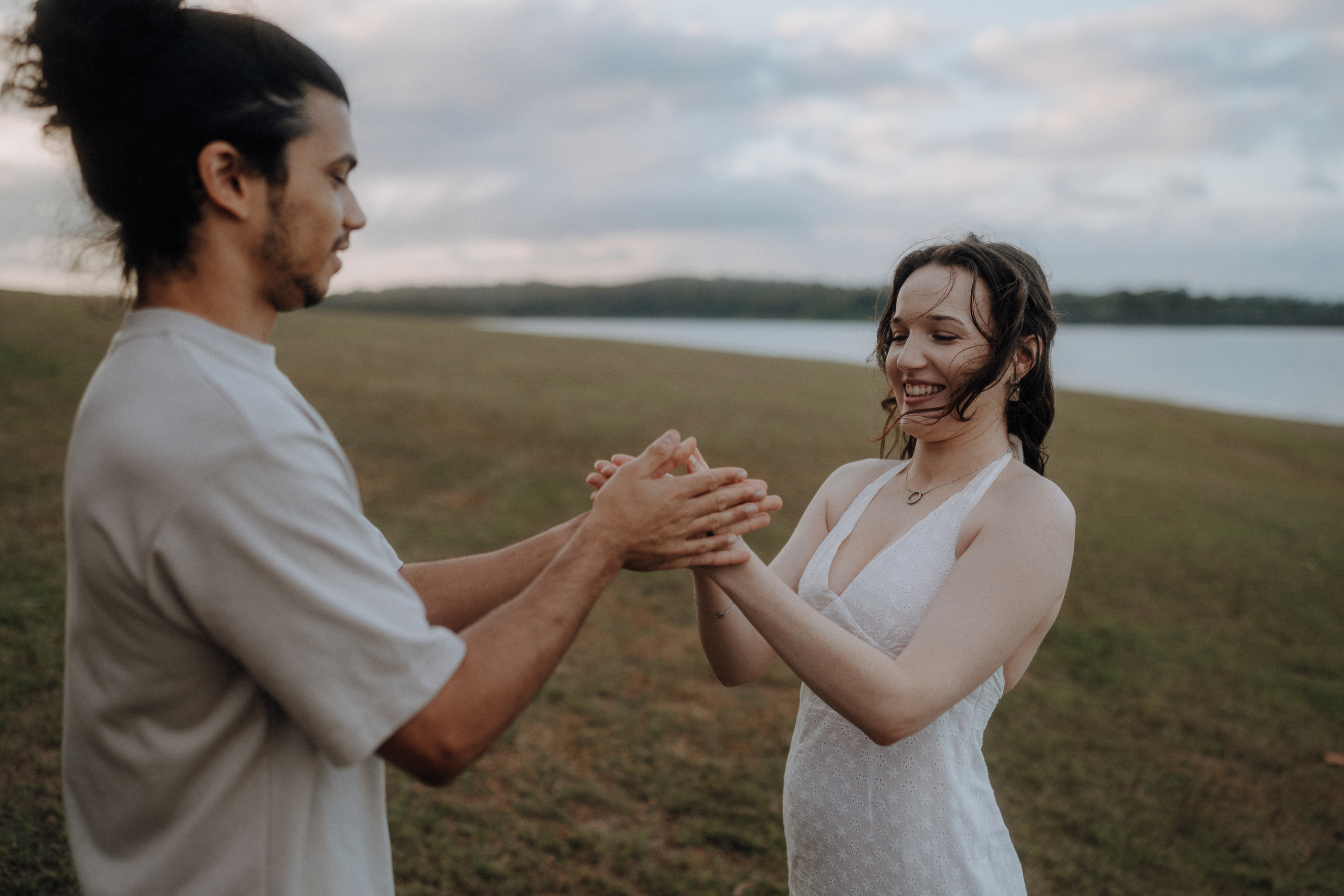 A man and woman standing outdoors on grass near water, facing each other and holding hands, both appearing relaxed and smiling.