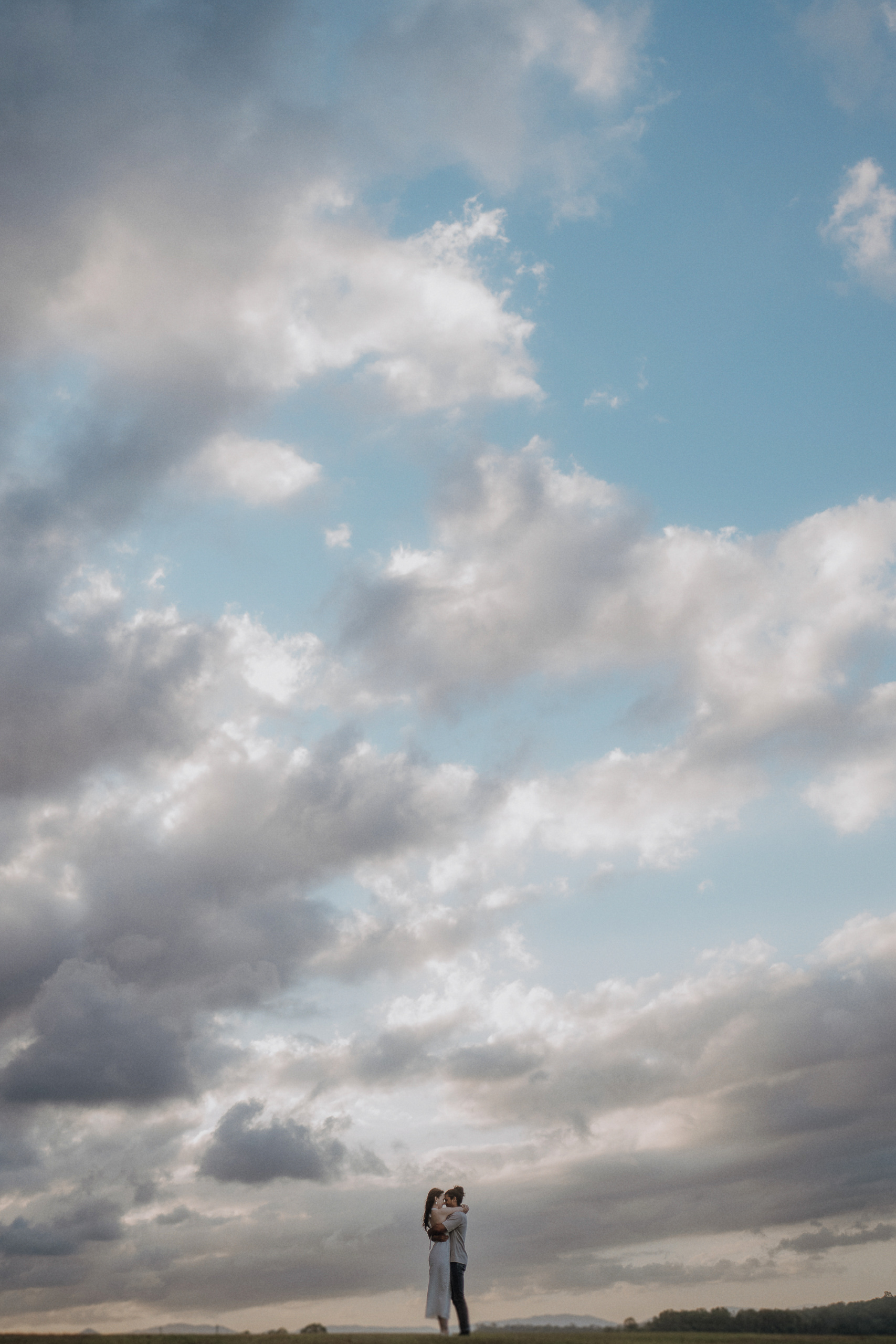 A couple stands close together in an open field under a vast, cloudy sky with patches of blue.