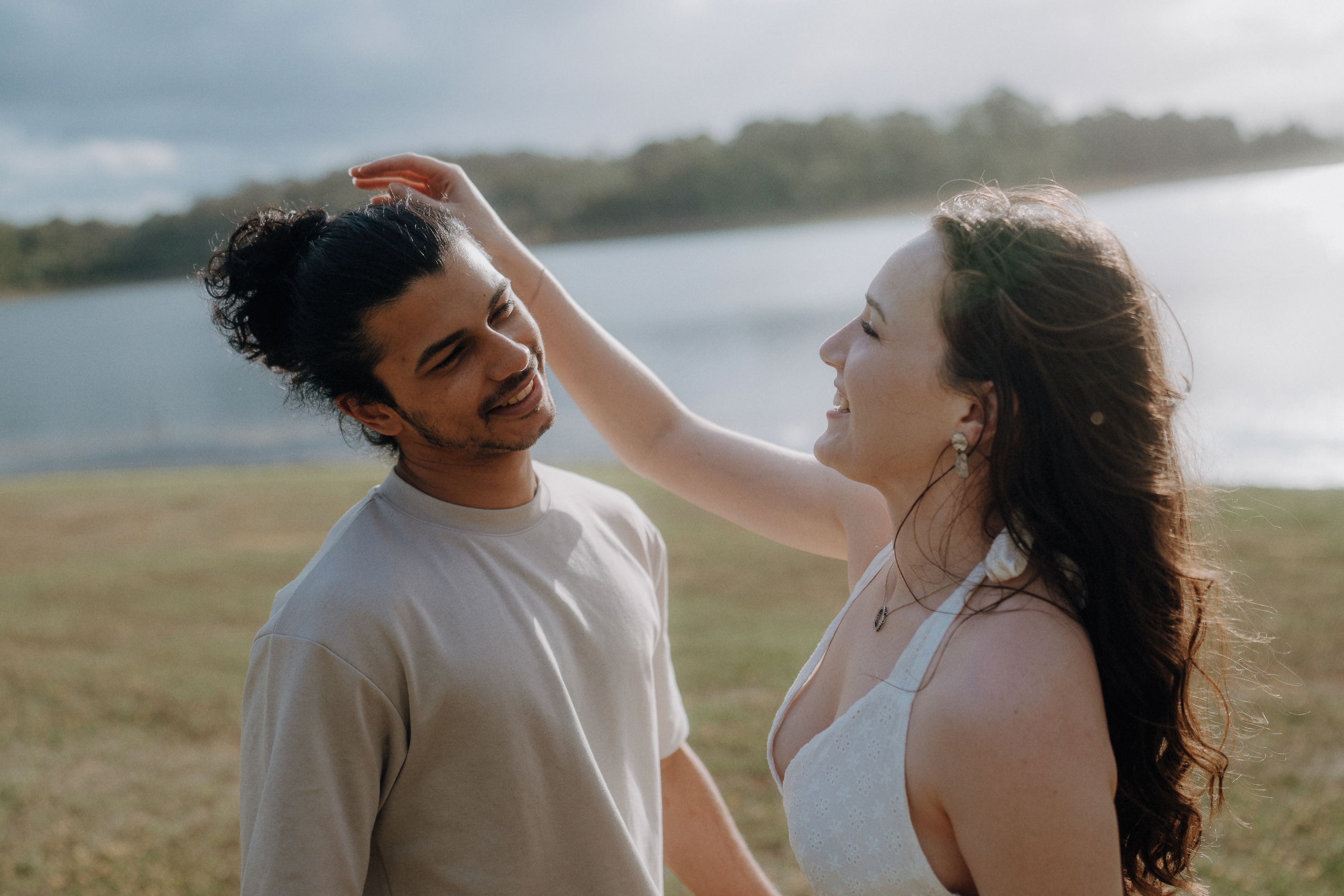 A woman smiles and lifts her hand toward a man's head as they stand together by a lake on a cloudy day.