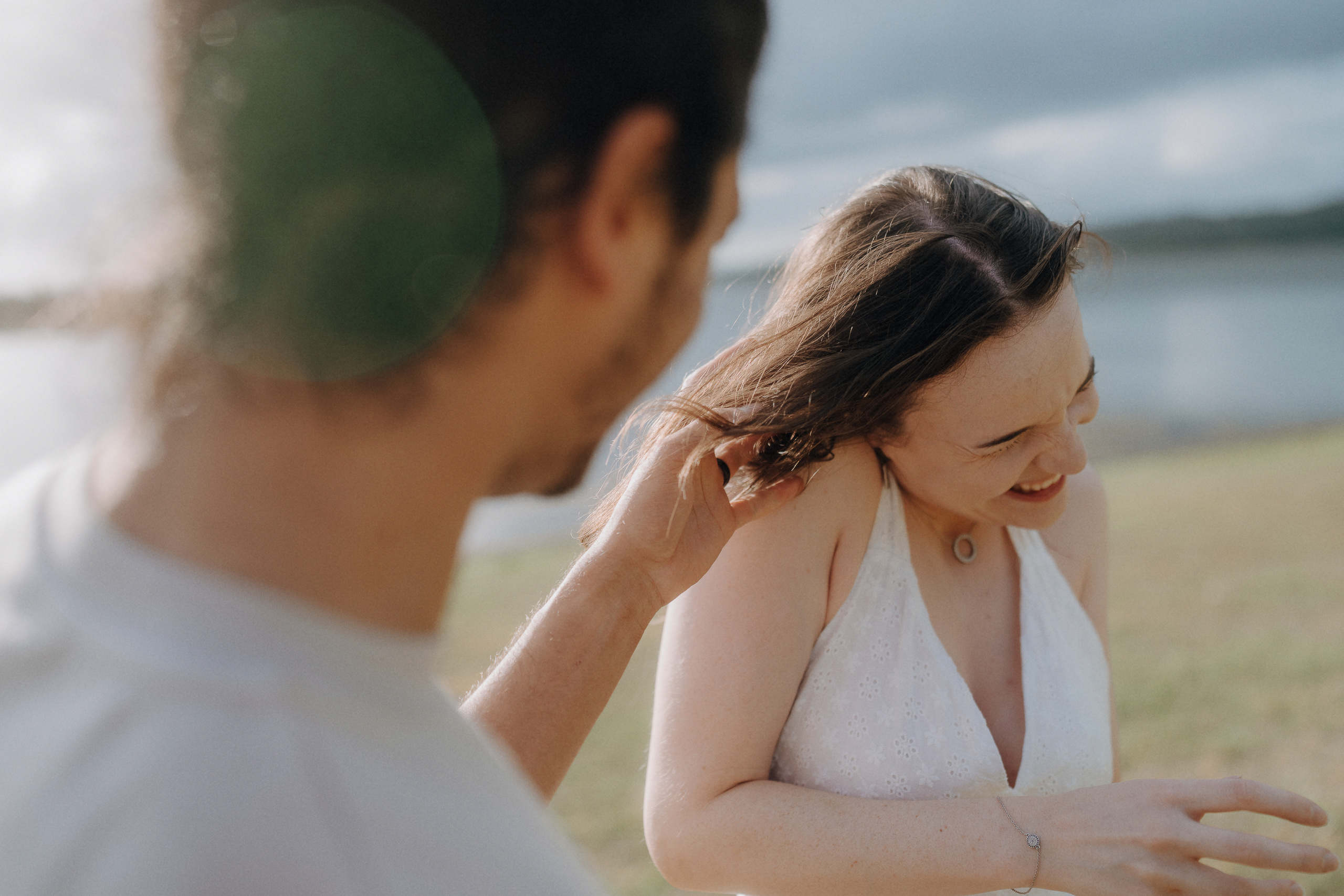 A man playfully tugs a woman's hair as she laughs and leans away, outdoors near water under a cloudy sky.