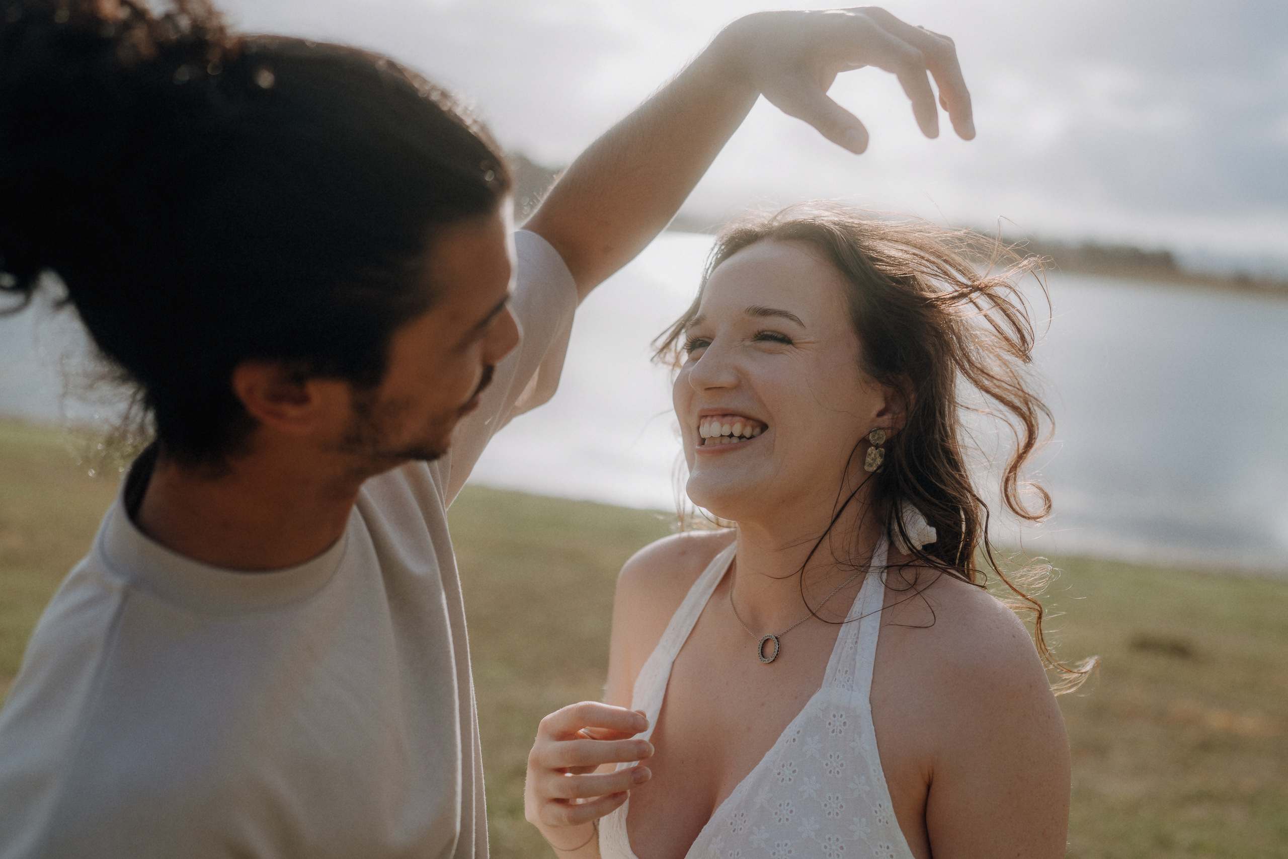 A man and woman stand outdoors by a body of water, smiling and laughing as he lifts his arm above her head.