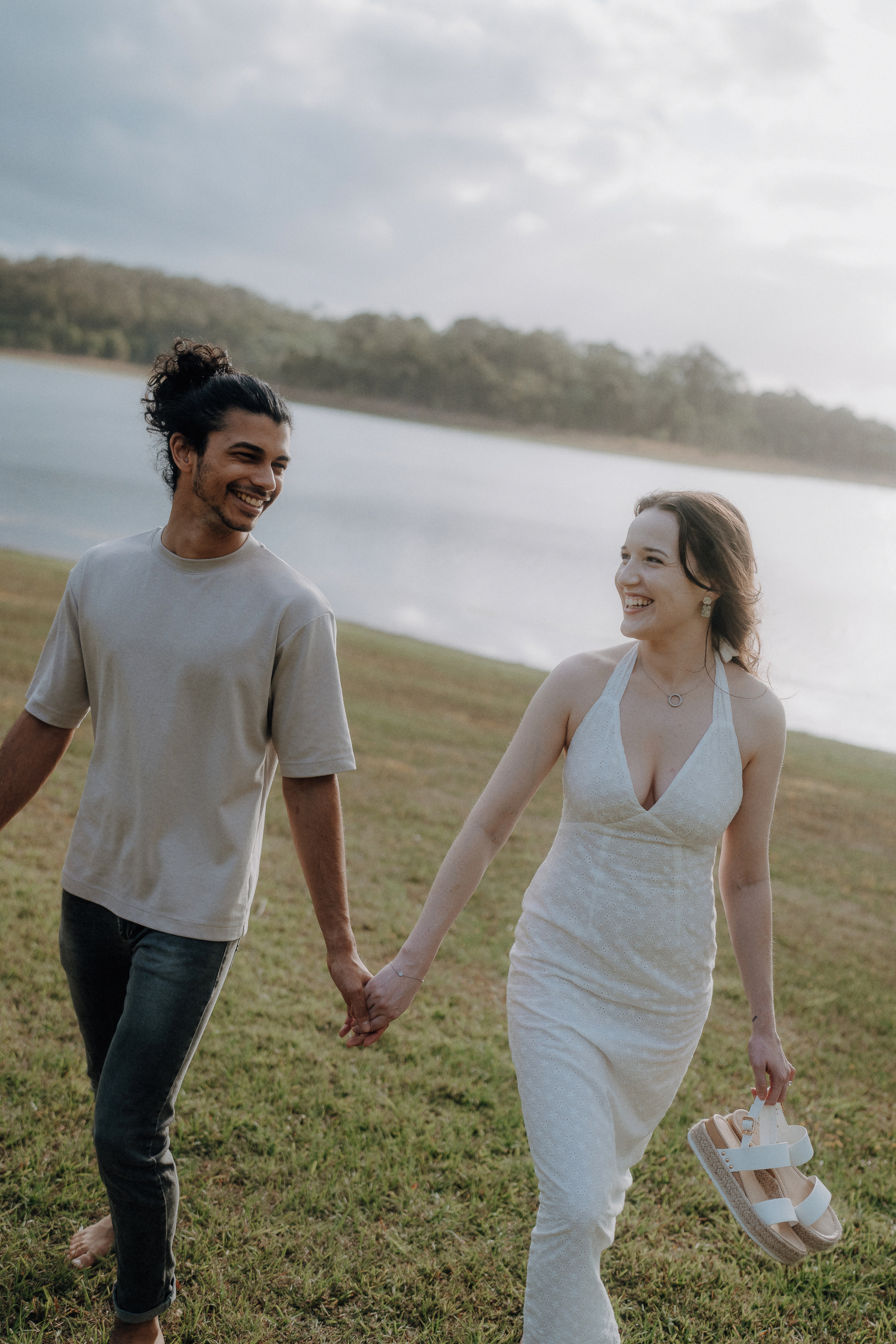 A smiling couple holding hands walks barefoot on grass beside a lake; the woman holds sandals and wears a white dress, while the man wears a light shirt and dark pants.