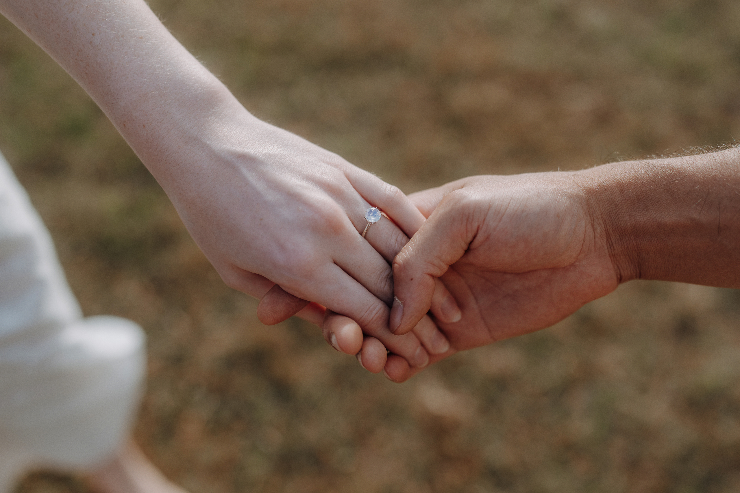 Close-up of two people holding hands outdoors, with one person wearing a diamond engagement ring.